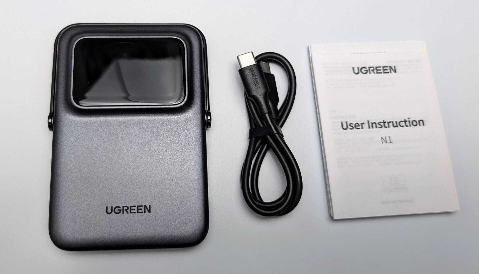 A UGREEN branded electronic power bank is shown with a sleek, metallic gray finish and a screen-like display at the top. Next to it lies a coiled black USB-C cable with the UGREEN logo. To the right, there's a folded UGREEN user instruction manual labeled "N1". The items are neatly arranged on a light background, highlighting their modern and minimalist design.