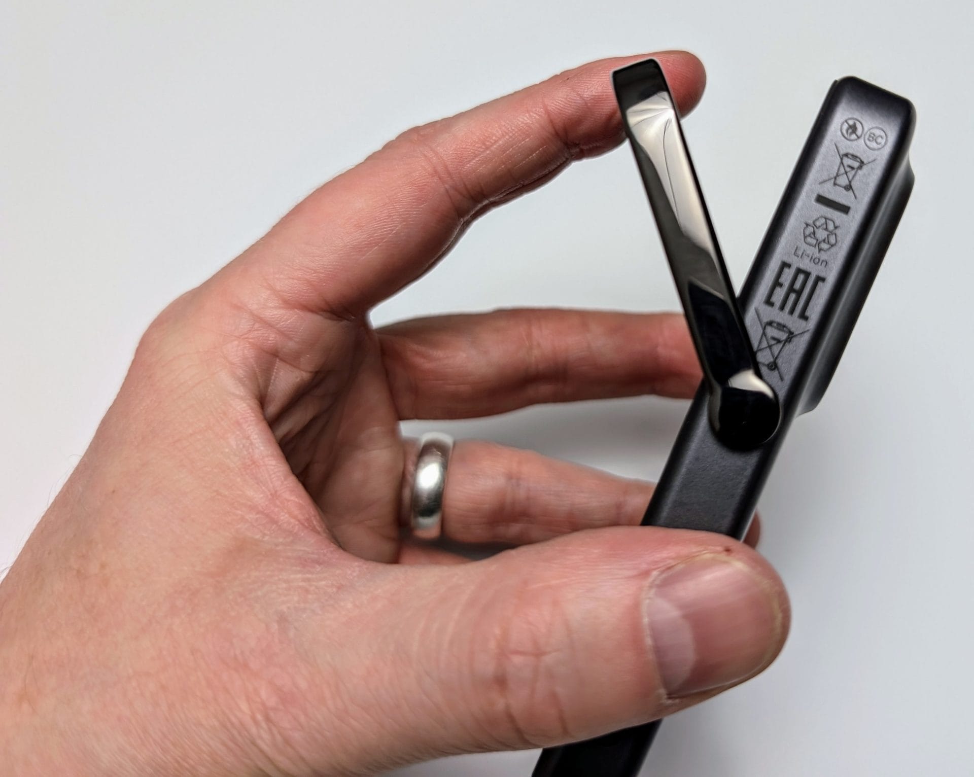 A close-up image of a hand holding a UGREEN Uno Magnetic Wireless Power device. The hand is gripping the device from the side, with the thumb and index finger. The device is grey with a foldable stand, currently in an open position. The device features various certification logos such as CE and EAC, and a lithium-ion battery symbol on its side. The background is a plain white surface.