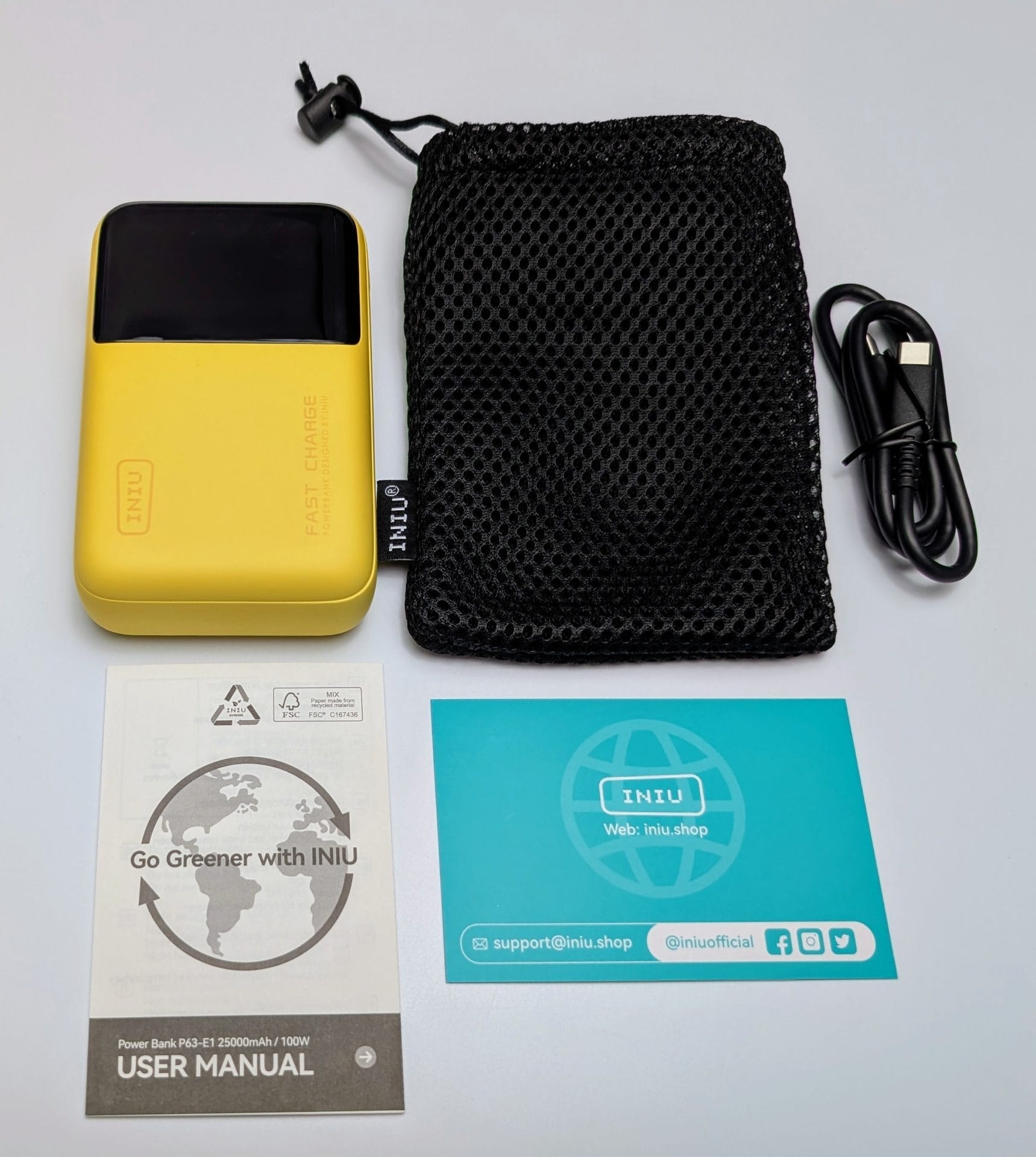 Contents of INIU Power Bank P63-E1 package laid out on a white surface. Includes a yellow INIU power bank labeled 'FAST CHARGE', a black mesh carrying pouch with the INIU logo, a coiled USB-C charging cable, a user manual with the text 'Go Greener with INIU' and details about the power bank, and a blue card with INIU branding, website, email support, and social media icons.