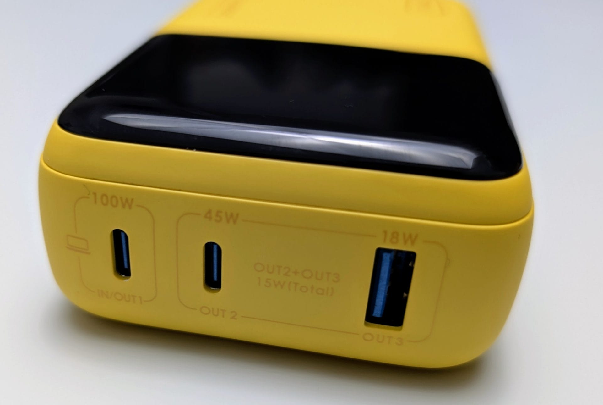 Close-up view of the bottom edge of a yellow INIU power bank. It features three ports: one labeled 'IN/OUT1' with a '100W' rating, one labeled 'OUT2' with a '45W' rating, and one labeled 'OUT3' with an '18W' rating. There's also a note indicating 'OUT2+OUT3 15W(Total)'