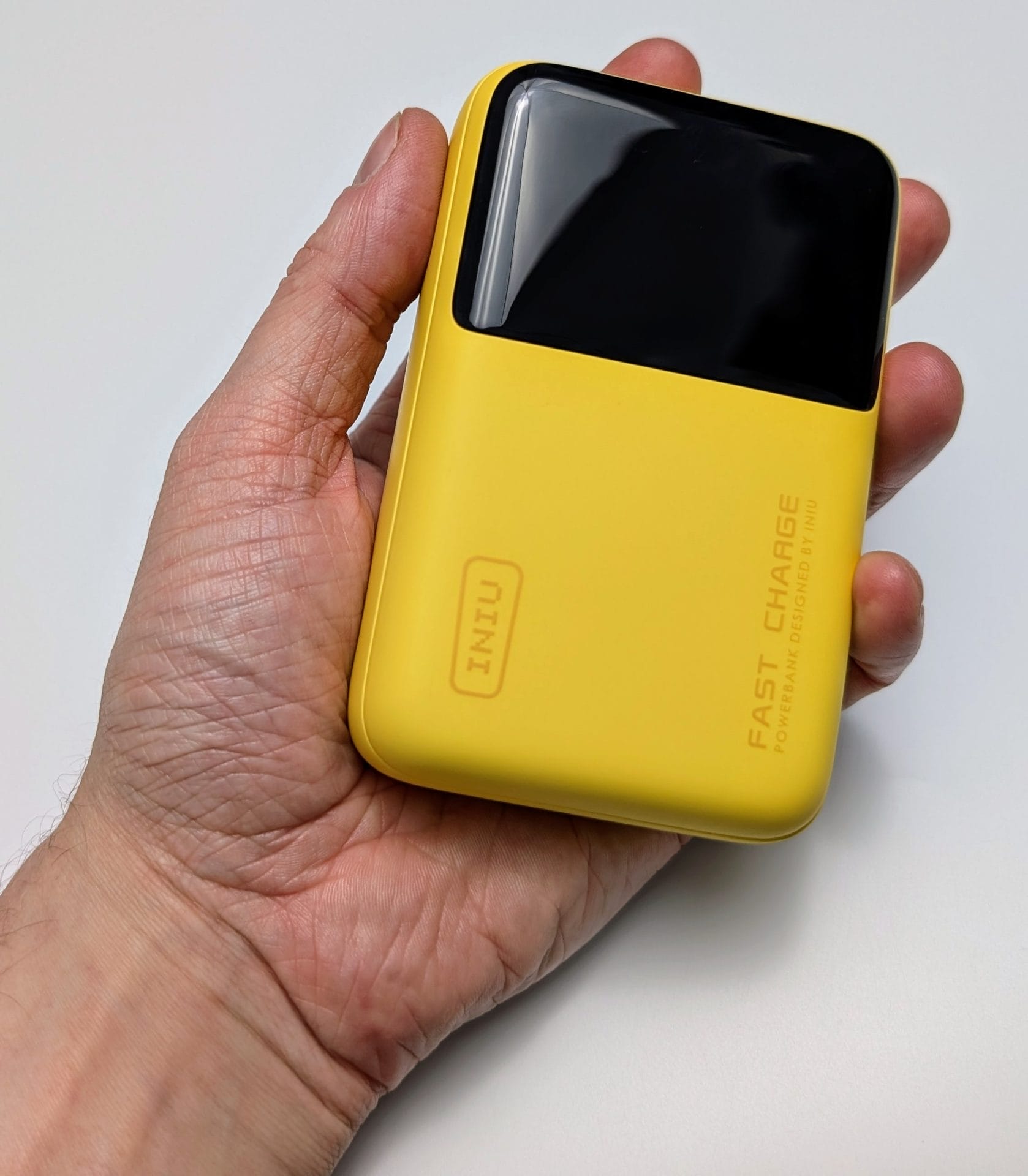 A hand holding a yellow INIU power bank with a black screen on top. The power bank is labeled 'FAST CHARGE' and 'POWER BANK CHARGE' on the side. The hand is positioned against a plain white background.