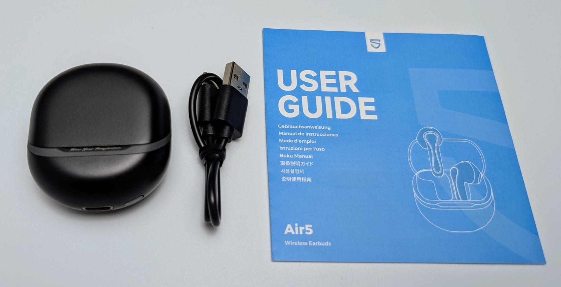 Image showing the accessories for SoundPEATS Air5 Wireless Earbuds. On the left, there is a sleek, black, oval-shaped charging case for the earbuds. Next to it, there is a black USB charging cable with a USB-A connector. On the right, there is a blue user guide with the title 'USER GUIDE' prominently displayed at the top. The guide includes translations of 'User Guide' in multiple languages such as German, Spanish, French, Italian, Japanese, Korean, and Chinese. Below the title, there's an illustration of the earbuds and their case. The model name 'Air5' and the description 'Wireless Earbuds' are printed at the bottom of the guide.