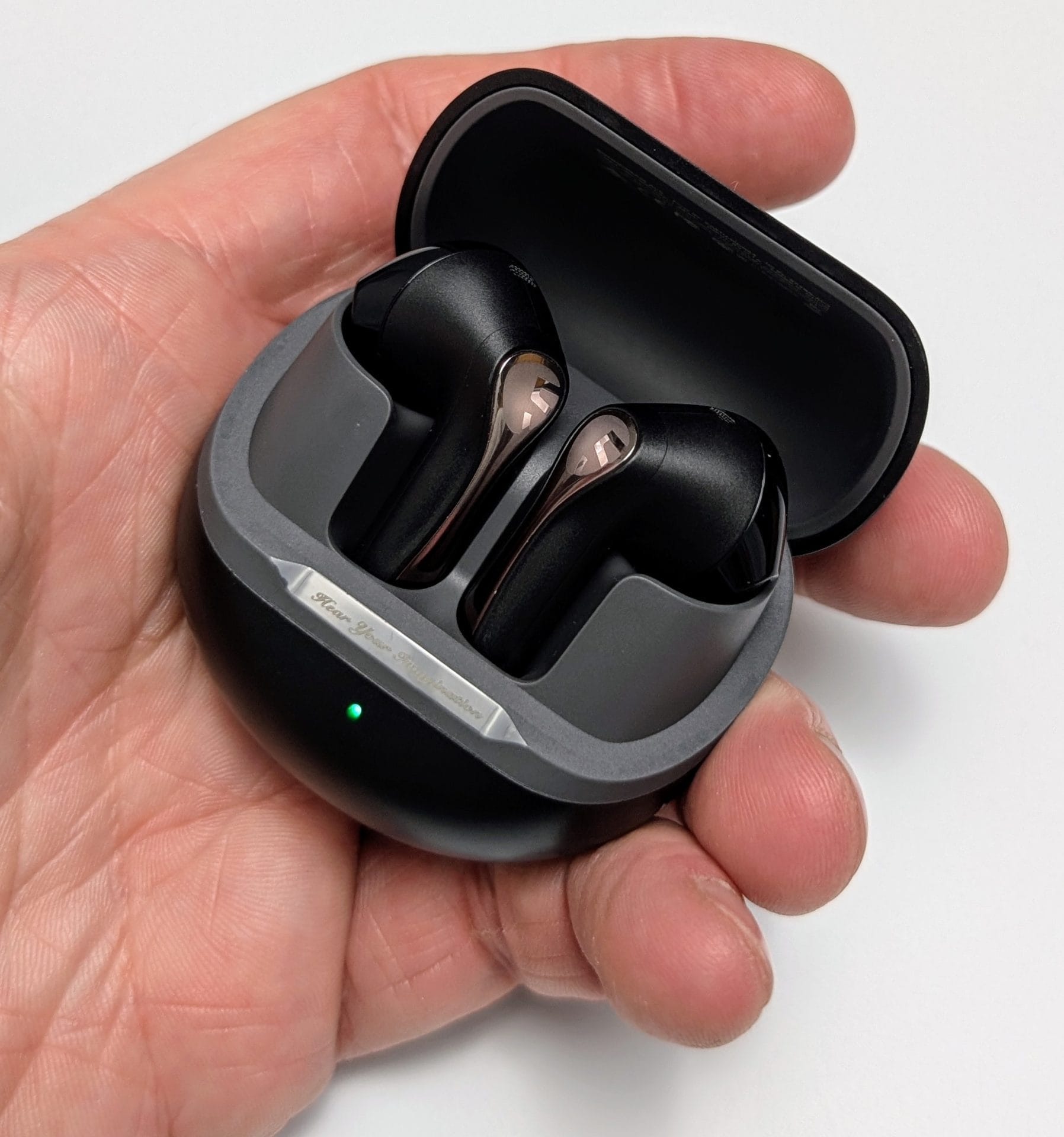 Image of a hand holding an open charging case for SoundPEATS Air5 Wireless Earbuds. The case is black with a sleek design, featuring a grey interior where the two earbuds are neatly placed. Each earbud is black with a metallic accent on the top. The case has a small green LED light indicating it is charging or fully charged. The exterior of the case has a metallic strip with the text 'For Your Imagination' engraved on it.