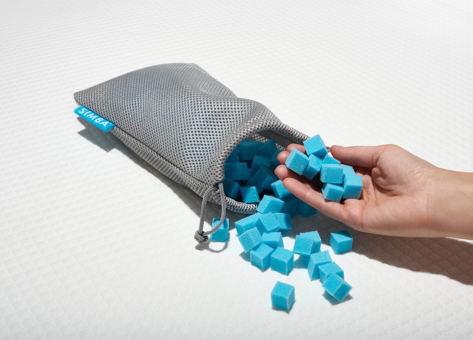 A hand holding several small blue foam cubes, with more cubes spilling out of an open gray mesh pouch featuring the Simba logo in blue. The pouch is placed on a textured white surface, likely a mattress or pillow, showcasing the adjustable foam filling used in Simba pillows for customizable comfort.