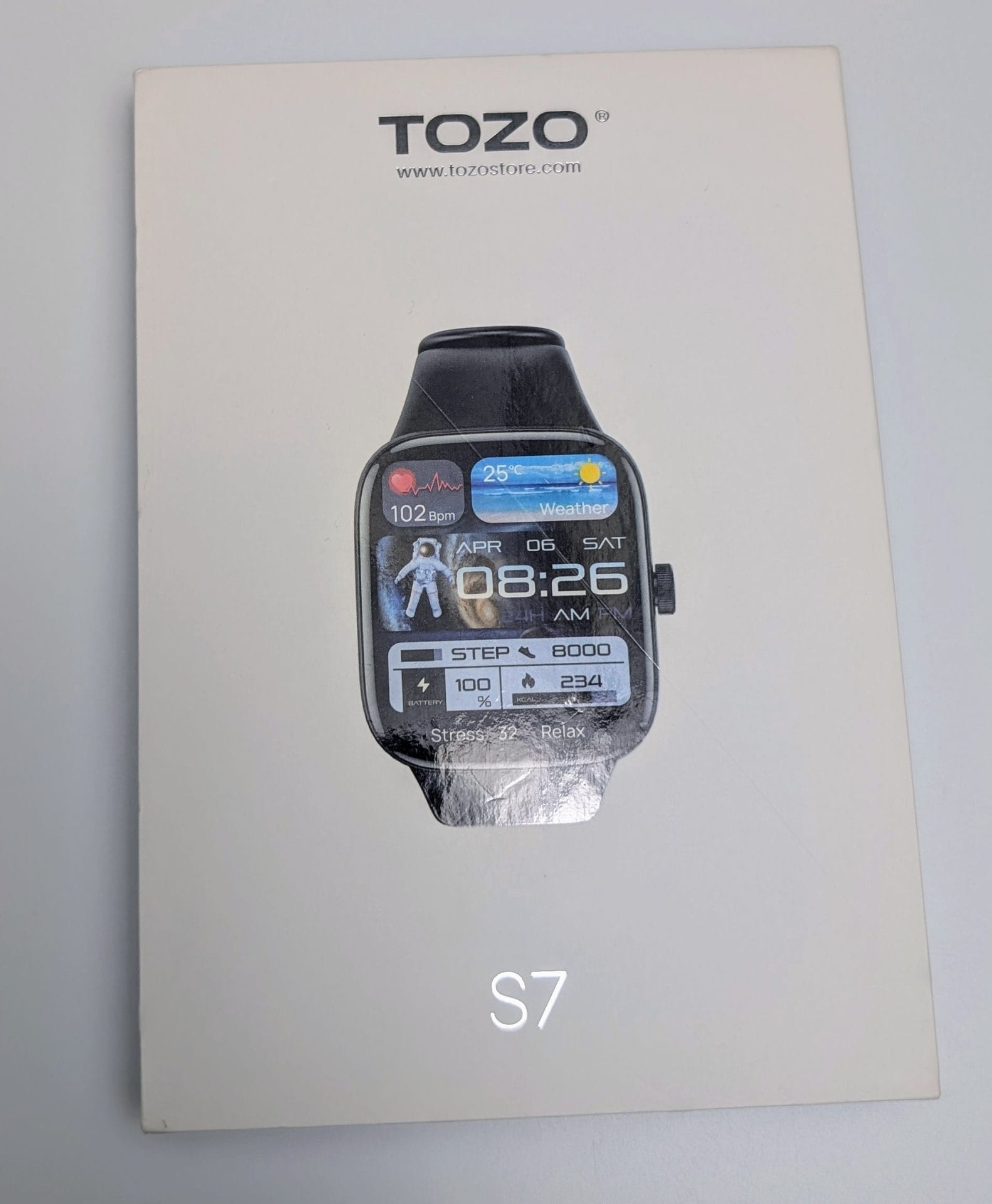 TOZO S7 Smartwatch A white box features the TOZO logo and website (www.tozostore.com) (www.tozostore.com) at the top. Below, an image of the TOZO S7 Smartwatch displays a rectangular black watch face with rounded corners. The screen shows the time (8:26 AM, Saturday, April 6), heart rate (102 bpm), weather, steps (8,000), battery (100%), and calories burned (234). An astronaut graphic is in the background. The text "Stress 32 Relax" is also visible. At the bottom, "S7" is written in large black letters.