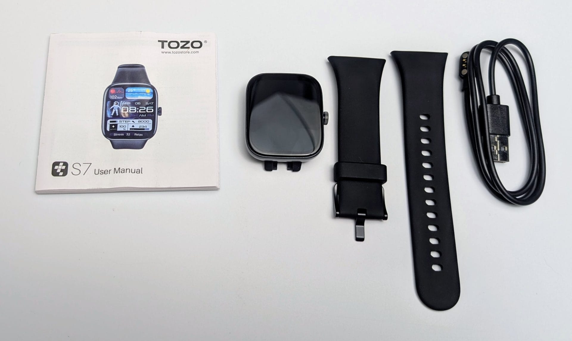A collection of items for the TOZO S7 Smartwatch is laid out on a white surface. From left to right: a user manual with the TOZO logo and "S7 User Manual" text, featuring an image of the watch (same as the previous image with time 8:26 AM, heart rate 102 bpm, steps 8,000, etc.); the S7 Smartwatch itself with a black rectangular face and rounded corners, screen off; a black silicone watch strap with a buckle and adjustment holes; and a black USB charging cable with a magnetic connector.
