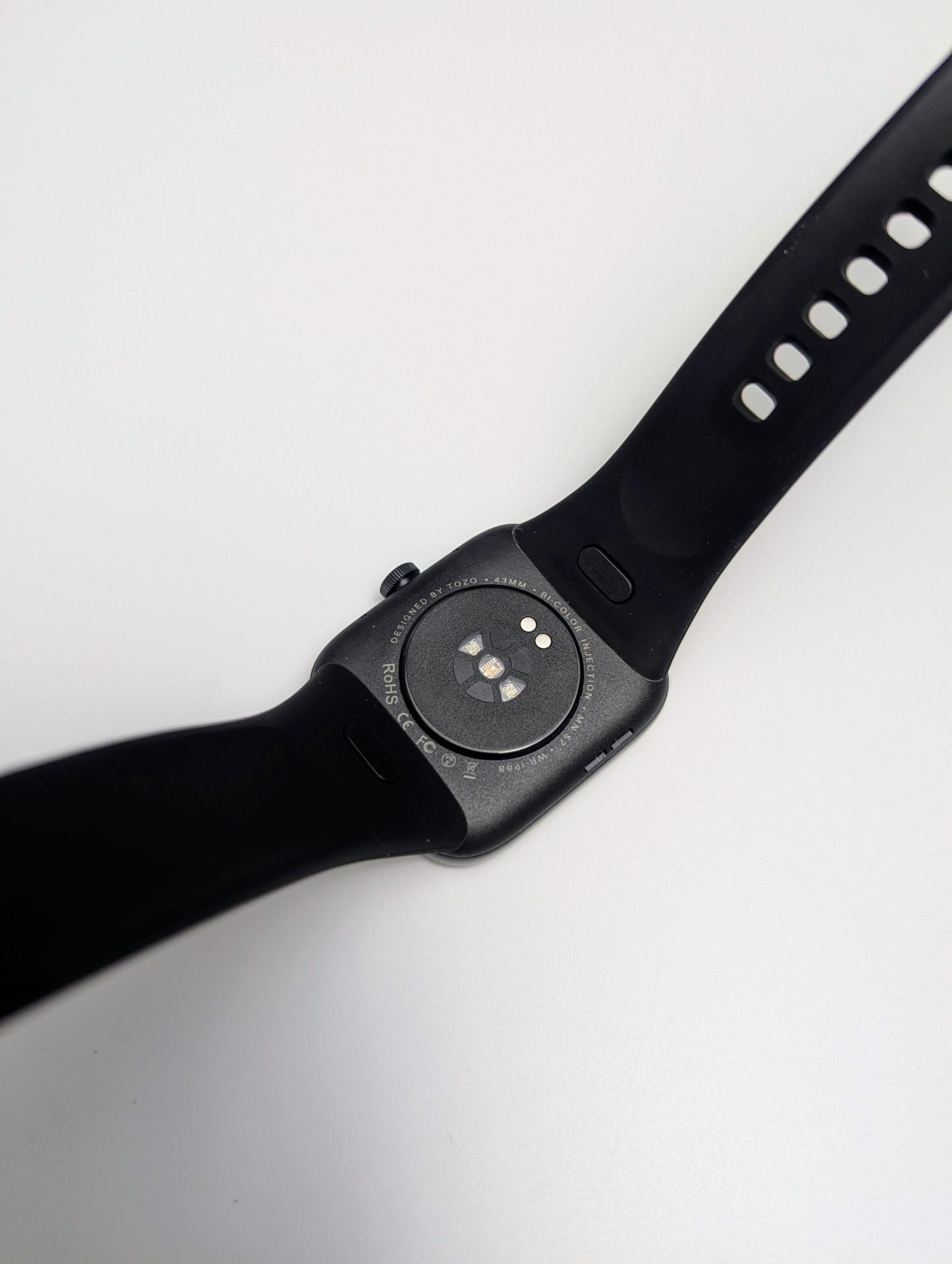 TOZO S7 Smartwatch The underside of the TOZO S7 Smartwatch is shown on a white surface. The black rectangular watch has a black silicone strap with adjustment holes. The back of the watch features a circular sensor area with multiple lights for heart rate monitoring, surrounded by text: "DESIGNED BY TOZO 5ATM WATERPROOF CE RoHS FCC." The strap is partially visible, showing its smooth texture and buckle attachment points.