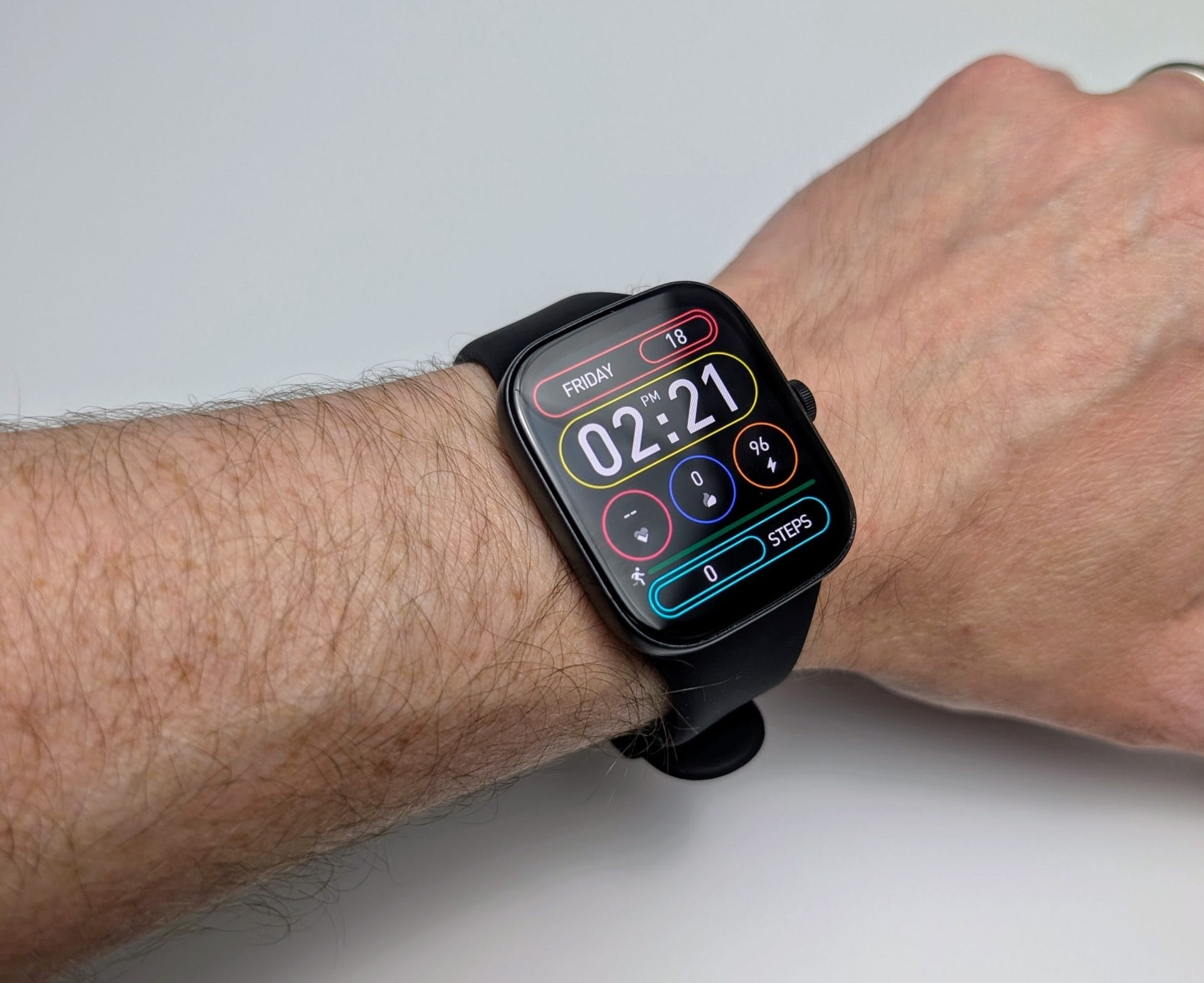 TOZO S7 Smartwatch A wrist wearing the TOZO S7 Smartwatch is shown against a white background. The black rectangular watch with rounded corners has a black silicone strap. The screen displays the time (02:21 PM, Friday), date (18), battery (0%), and steps (0), with icons for heart rate and other functions. The strap is secured around the wrist, showing its smooth texture and fit on a hairy forearm.