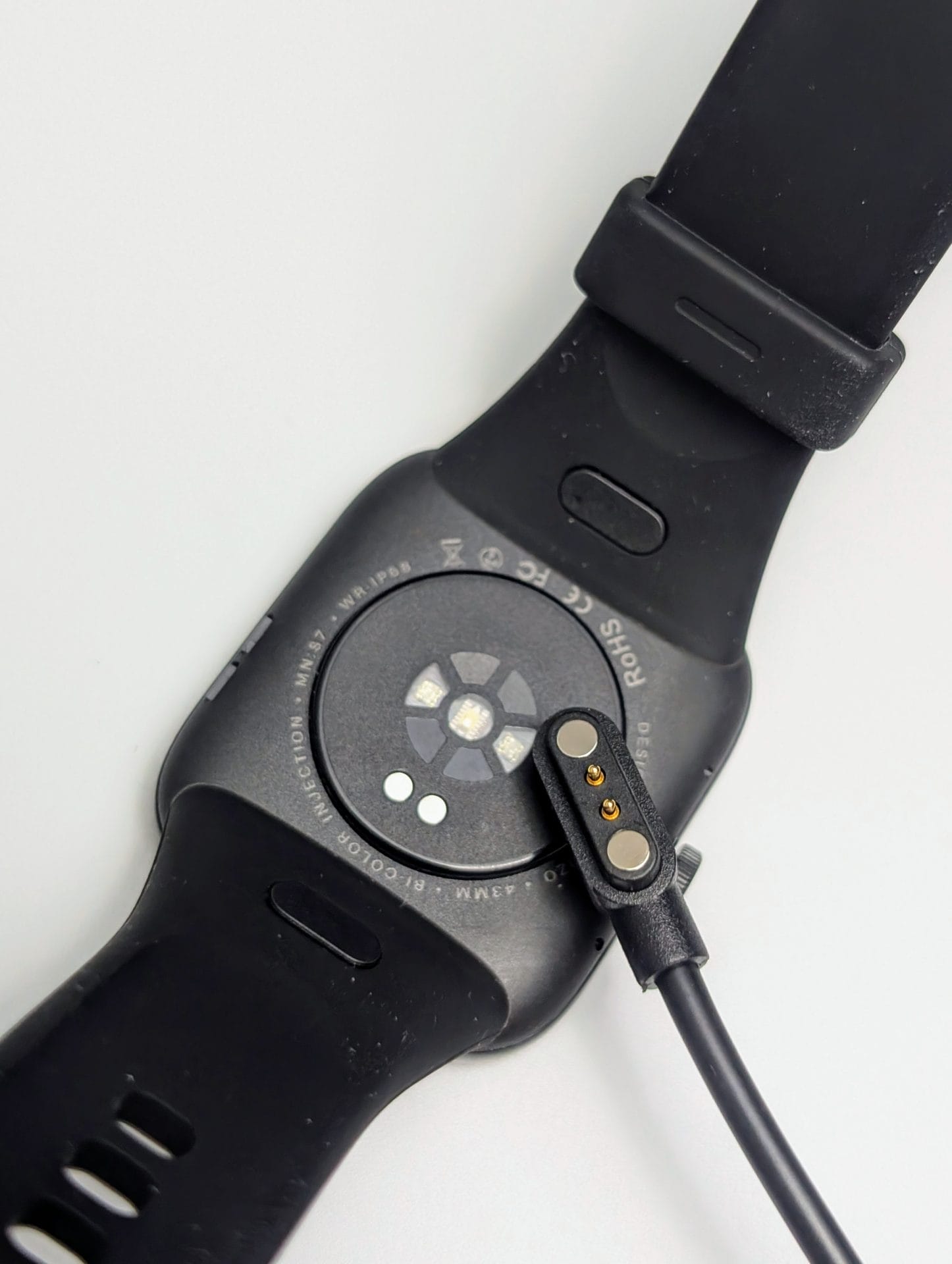 TOZO S7 Smartwatch The underside of the TOZO S7 Smartwatch is shown on a white surface, connected to its charging cable. The black rectangular watch has a black silicone strap with adjustment holes. The back features a circular sensor area with lights for heart rate monitoring, surrounded by text: "DESIGNED BY TOZO 5ATM WATERPROOF CE RoHS FCC." A magnetic charging connector with two gold pins is attached to the sensor area, with a black cable extending from it. The strap shows its smooth texture and buckle attachment points.