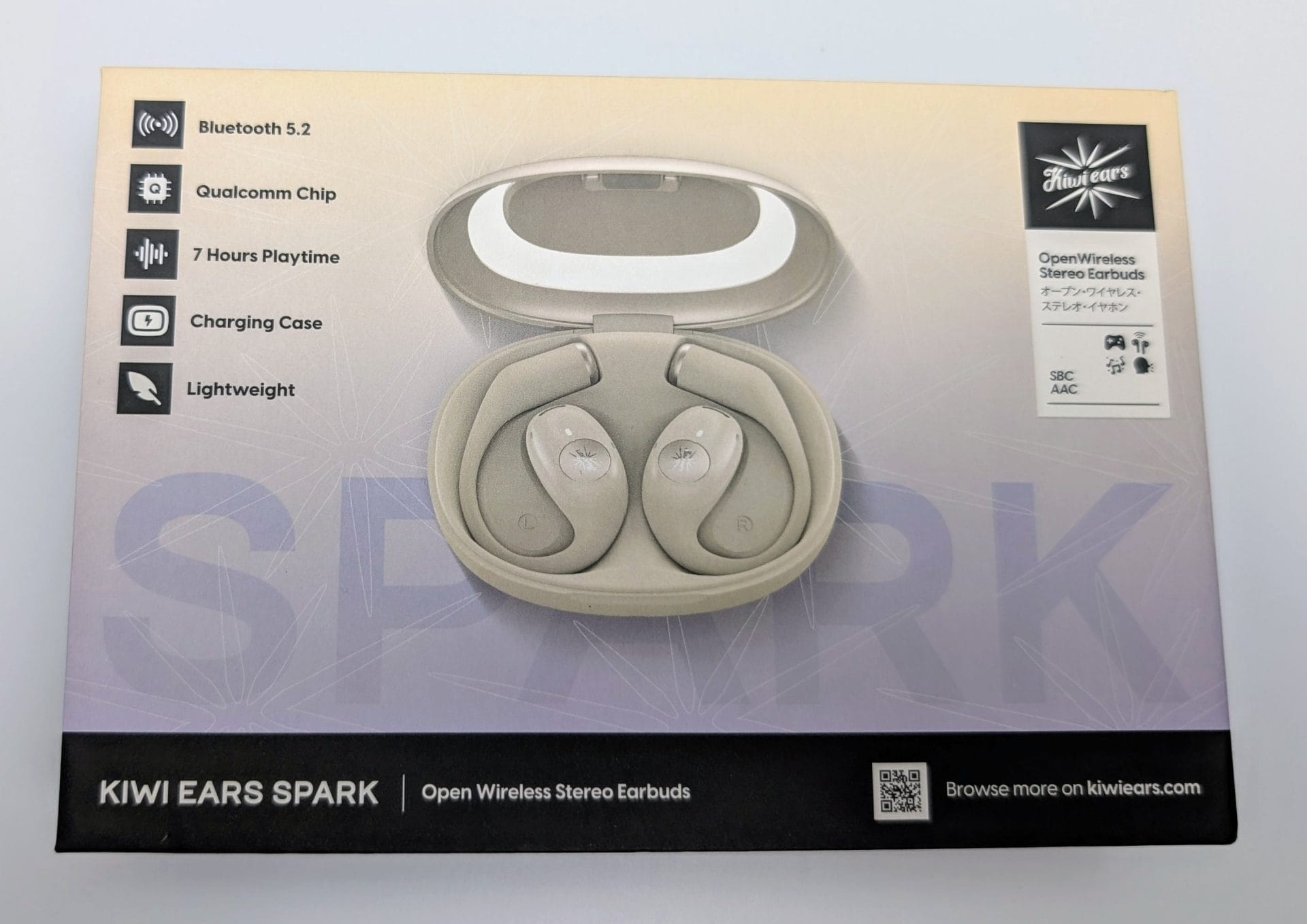A beige and black box for Kiwi Ears Spark Open Wireless Stereo Earbuds. The center features an image of two white earbuds in an open charging case. On the left, icons and text highlight features: Bluetooth 5.2, Qualcomm Chip, 7 hours playtime, charging case, and lightweight design. On the right, text reads "OPEN WIRELESS STEREO EARBUDS" in English and Japanese, with "SBC AAC" and icons for gaming and music. The bottom has the text "KIWI EARS SPARK | Open Wireless Stereo Earbuds" and a QR code with "Browse more on kiwiears.com." The top right corner has the Kiwi Ears logo.