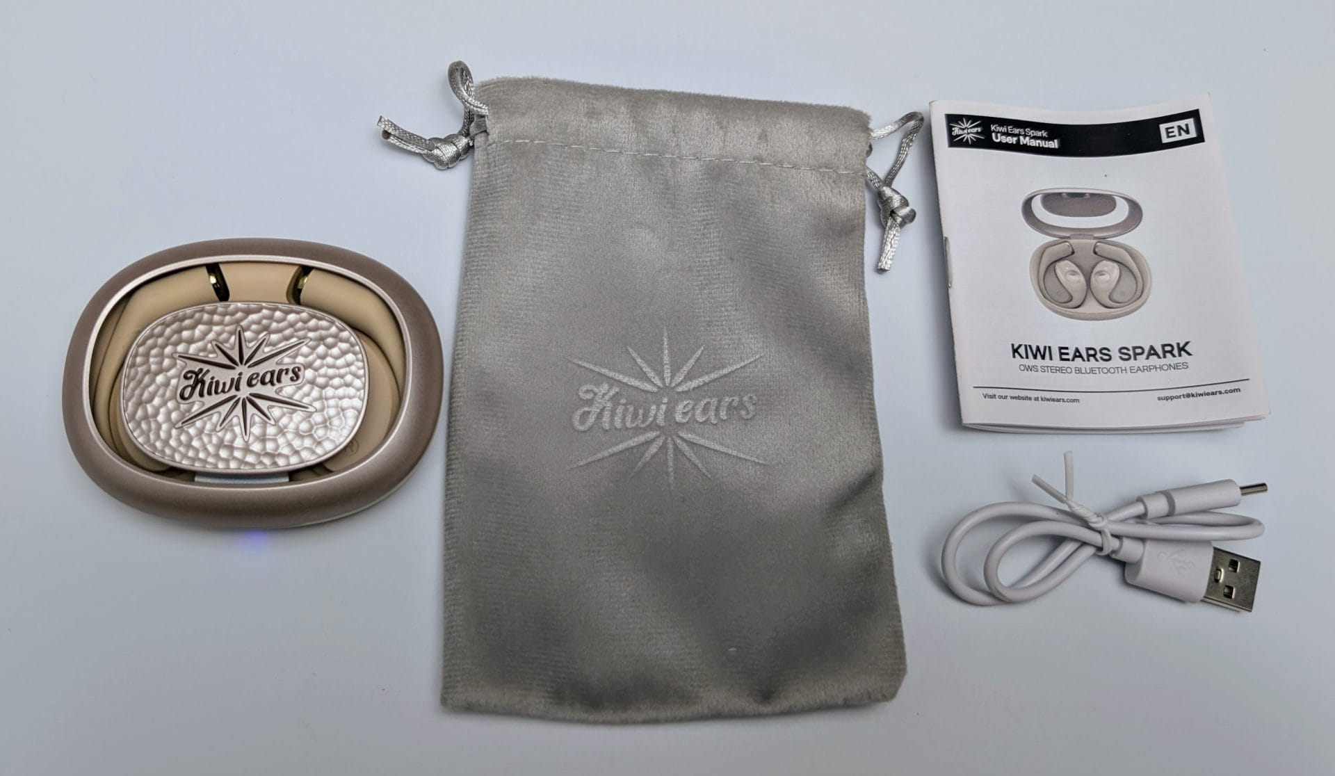 Kiwi Ears Spark A collection of items from a Kiwi Ears Spark package on a white background. On the left, a beige charging case with a textured silver lid featuring the "Kiwi ears" logo and a starburst design. In the center, a gray drawstring pouch with "Kiwi ears" and a starburst logo embossed on it. On the right, a white user manual for "KIWI EARS SPARK OWS Stereo Bluetooth Earphones" with an image of the earbuds in their case, labeled "EN" for English. Below the manual, a white USB charging cable with a USB-A to USB-C connector.