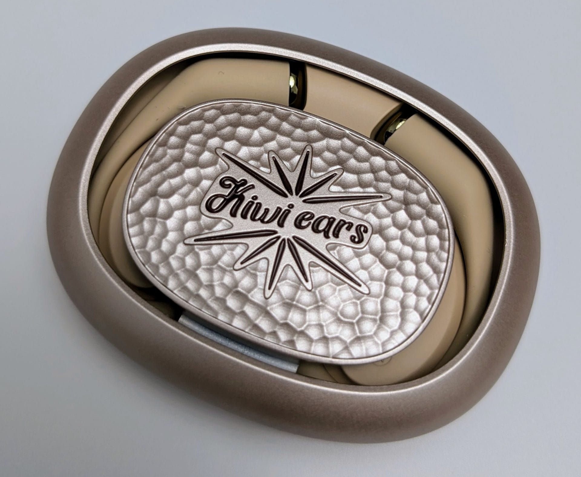 A closed beige charging case for Kiwi Ears Spark earbuds on a white background. The case has a textured silver lid with the "Kiwi ears" logo and a starburst design embossed on it. The case is oval-shaped with a smooth, glossy finish on the outer rim.