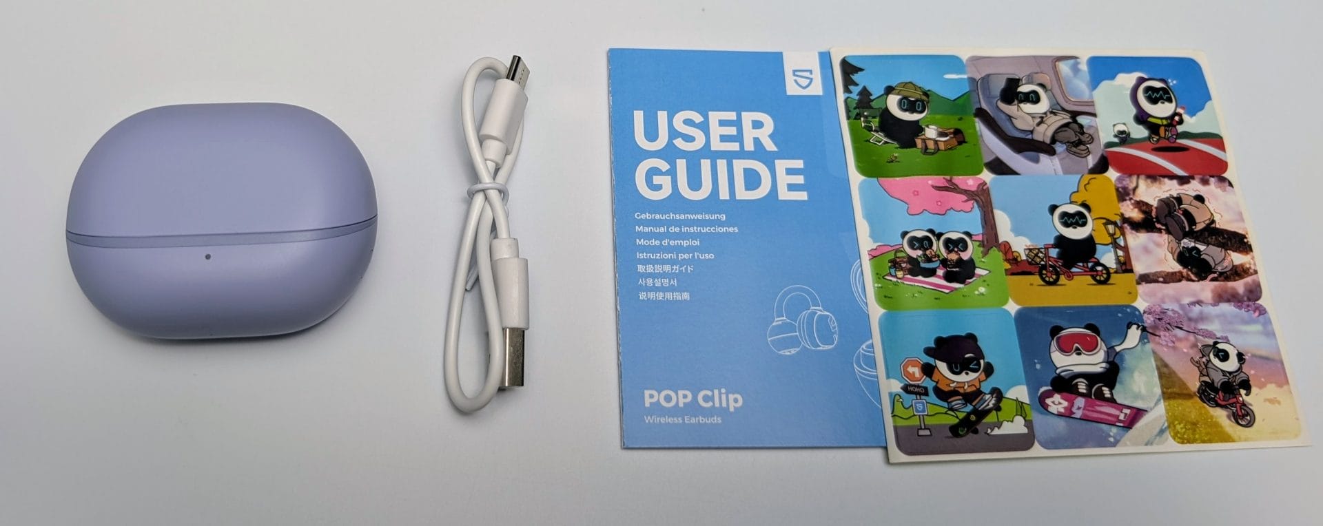 SoundPEATS POP Clip Earbuds A light purple charging case for SoundPEATS POP Clip Earbuds is placed next to a coiled white USB-C charging cable. To the right, a blue user guide booklet with "USER GUIDE" and "POP Clip Wireless Earbuds" text in white is accompanied by a sheet of colorful stickers featuring cartoon pandas in various playful scenes, such as skateboarding and cycling.