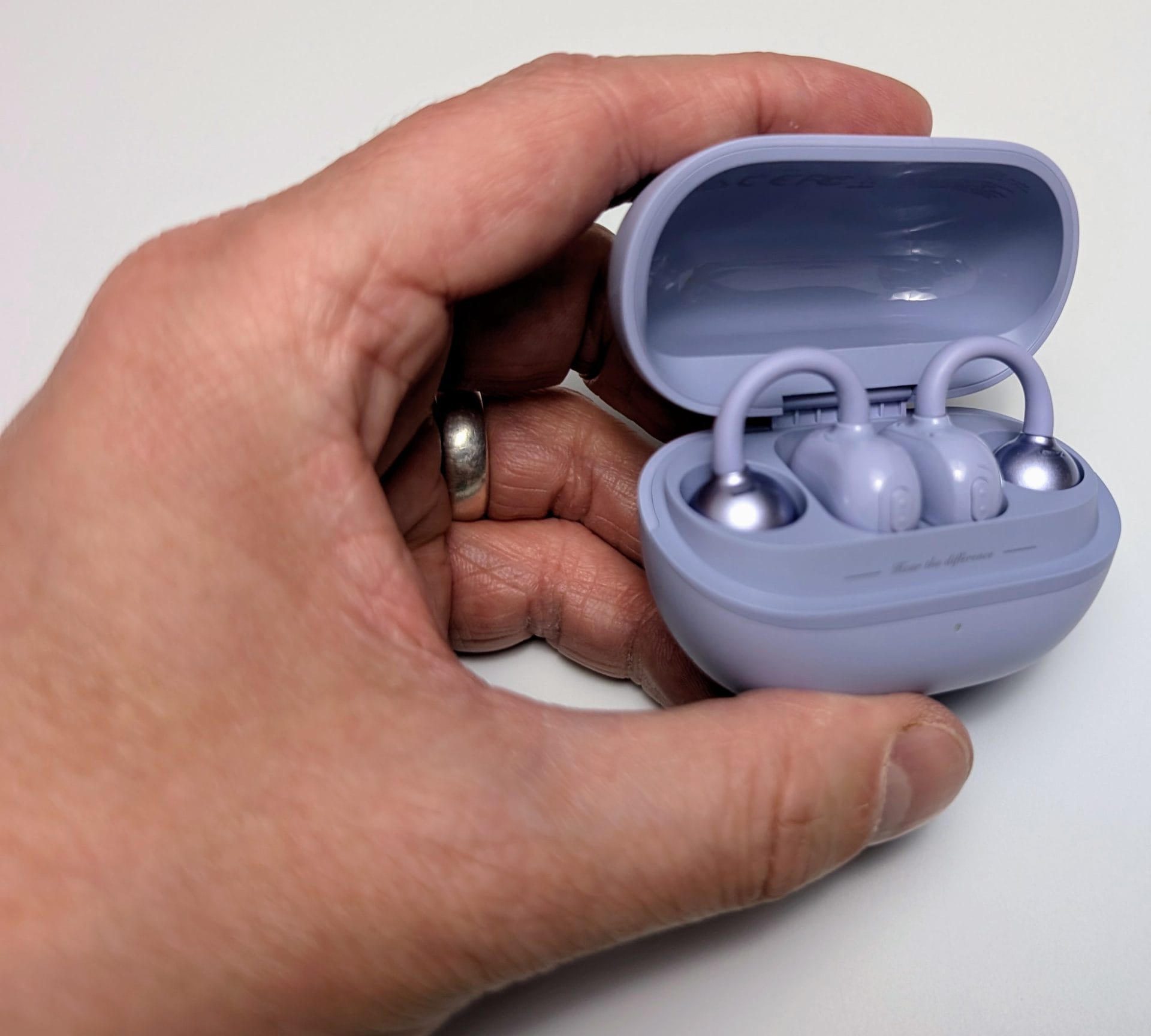 A hand with a silver ring holds an open light purple charging case for SoundPEATS POP Clip Earbuds, revealing two earbuds inside. The earbuds are light purple with silver tips and clip-style hooks, nestled in their charging slots within the case.