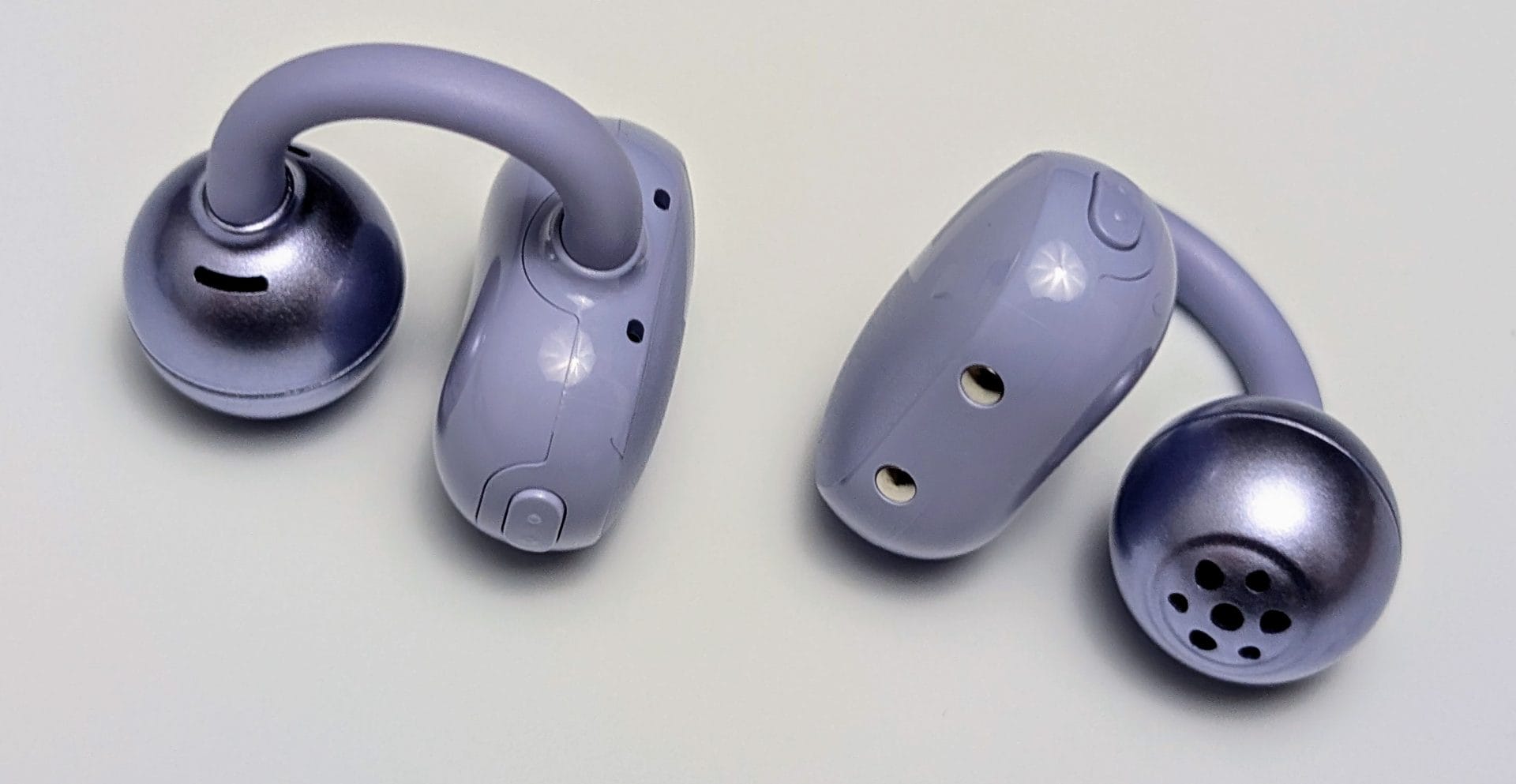 SoundPEATS POP Clip Earbuds Two SoundPEATS POP Clip Earbuds in light purple with silver tips are shown from different angles. Each earbud features a clip-style hook, with one displaying a perforated speaker grille and the other showing small ventilation holes and buttons on the side.