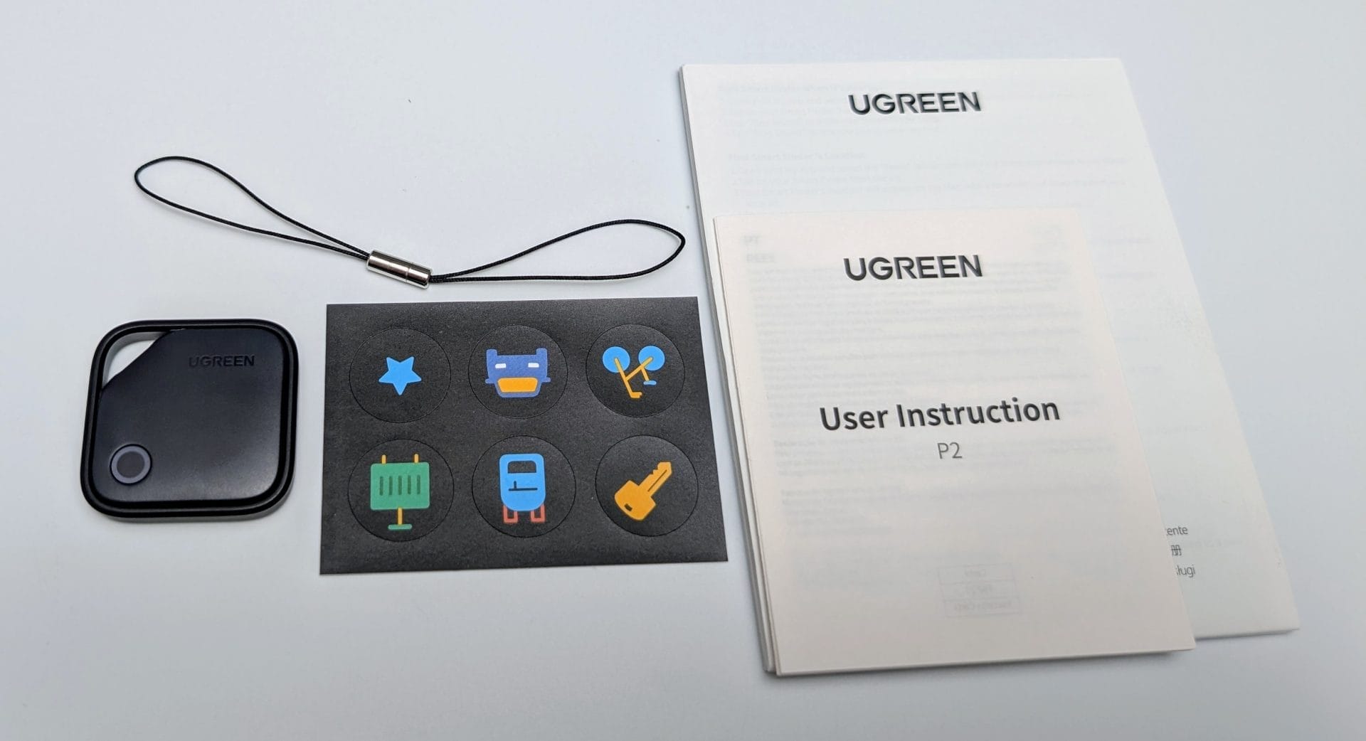 UGREEN FineTrack Smart Finder A collection of items from a UGREEN Smart Finder package on a white background. On the left, a black UGREEN Smart Finder device with a circular button and the UGREEN logo. Next to it, a black lanyard with a metal clasp. In the center, a black card with six colorful stickers: a blue star, a yellow laptop, blue headphones, a green wallet, a blue train, and a yellow key. On the right, two white UGREEN user instruction manuals, one labeled "User Instruction P2" with additional text in smaller font.