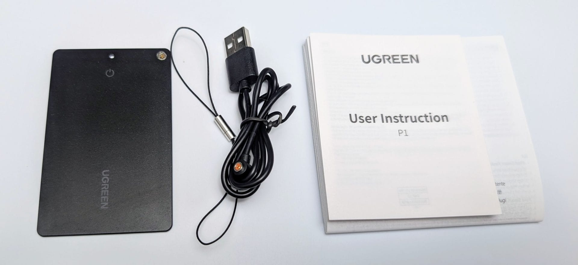 UGREEN FineTrack Slim Smart Finder A UGREEN FineTrack Slim Smart Finder package contents laid out on a gradient blue background: a thin black rectangular device with a power button and small hole, a black lanyard with a metal clip, a black USB charging cable, and a user instruction manual labeled "UGREEN User instruction P1."