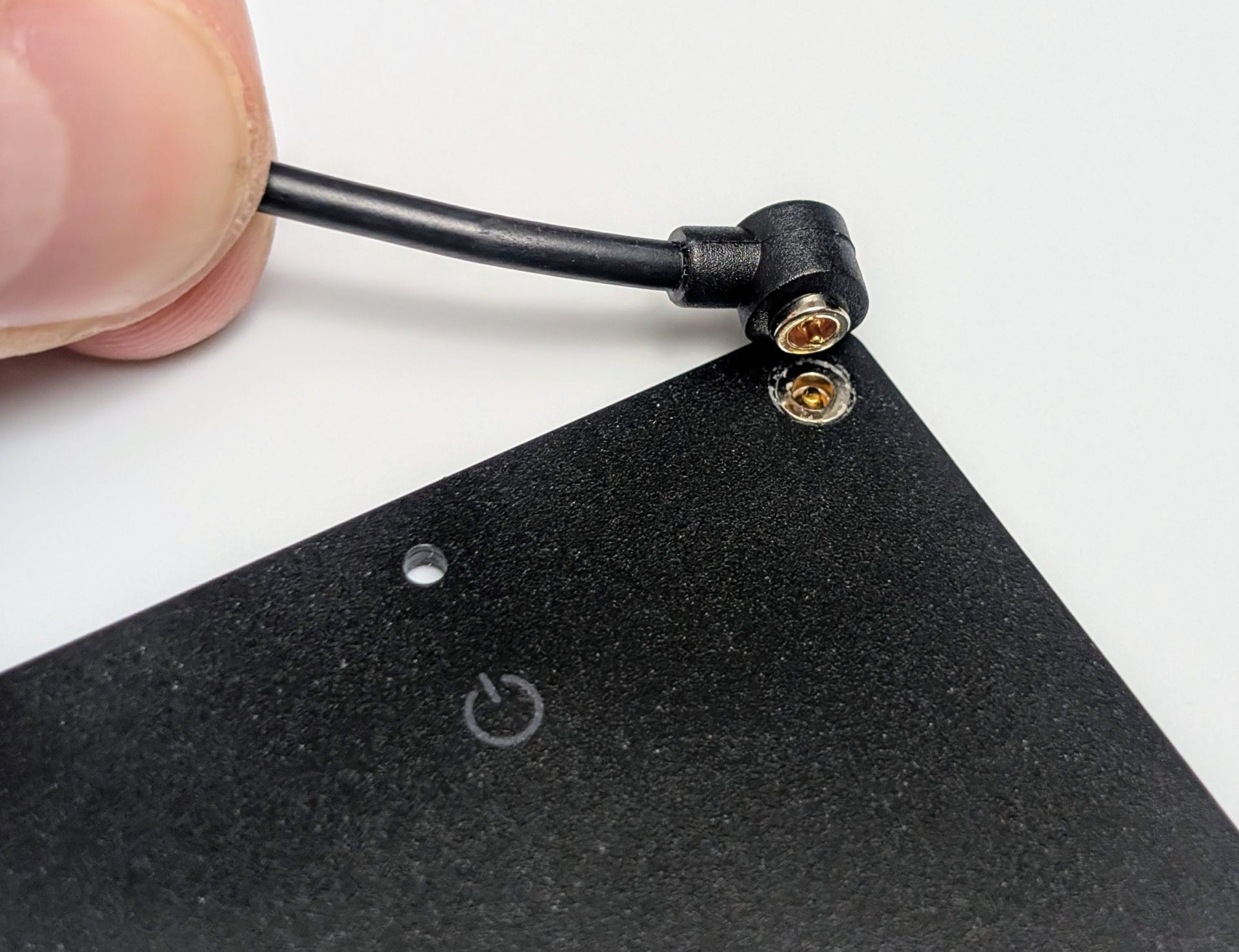 A close-up of a hand holding a black charging cable with a right-angle connector, aligning it with the charging port on a UGREEN FineTrack Slim Smart Finder, a thin black rectangular device with a power button and a small hole visible on its surface, against a white background.