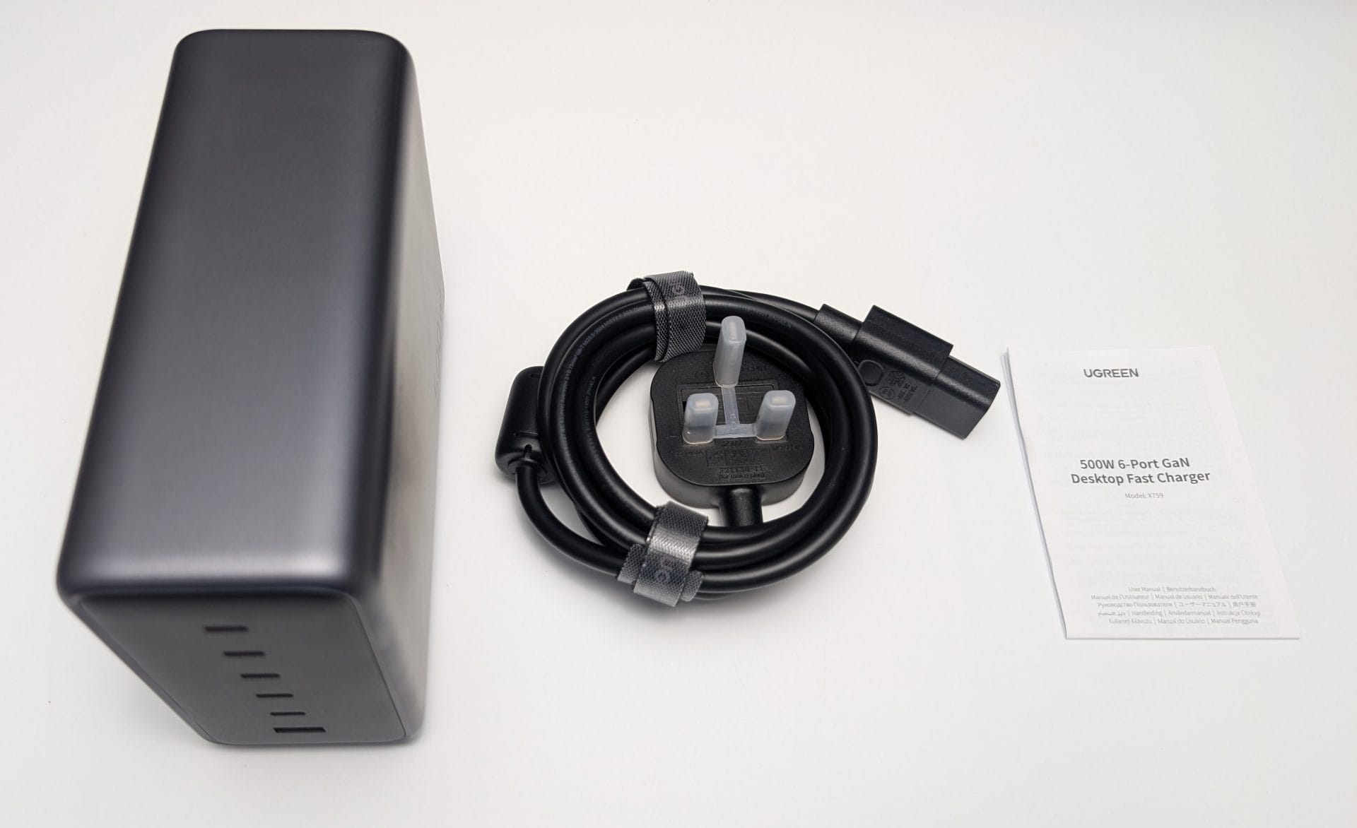 The unboxed UGREEN Nexode 500W 6-Port GaN Desktop Charger (Model: 15796) in grey, placed on a white surface. It features multiple ports on one side. Next to it is a coiled 2-metre black power cord with a UK plug, secured with a strap. A folded instruction manual labelled "UGREEN 500W 6-Port GaN Desktop Charger" is also included. The setup is photographed on 31 May 2025 at 09:57 AM BST.