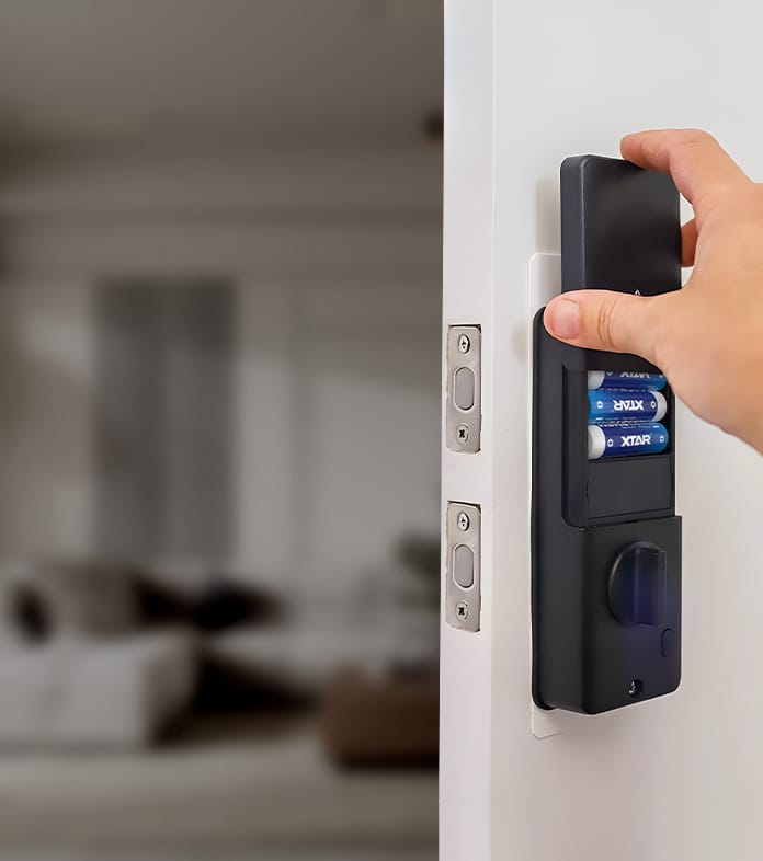 XTAR 1.5V AA LR 3000mAh USB-C Lithium Batteries A hand inserting four XTAR AA batteries into the battery compartment of a black smart lock mounted on a white door. The background shows a blurred indoor setting with a sofa and furniture. The lock is positioned next to a traditional door latch.