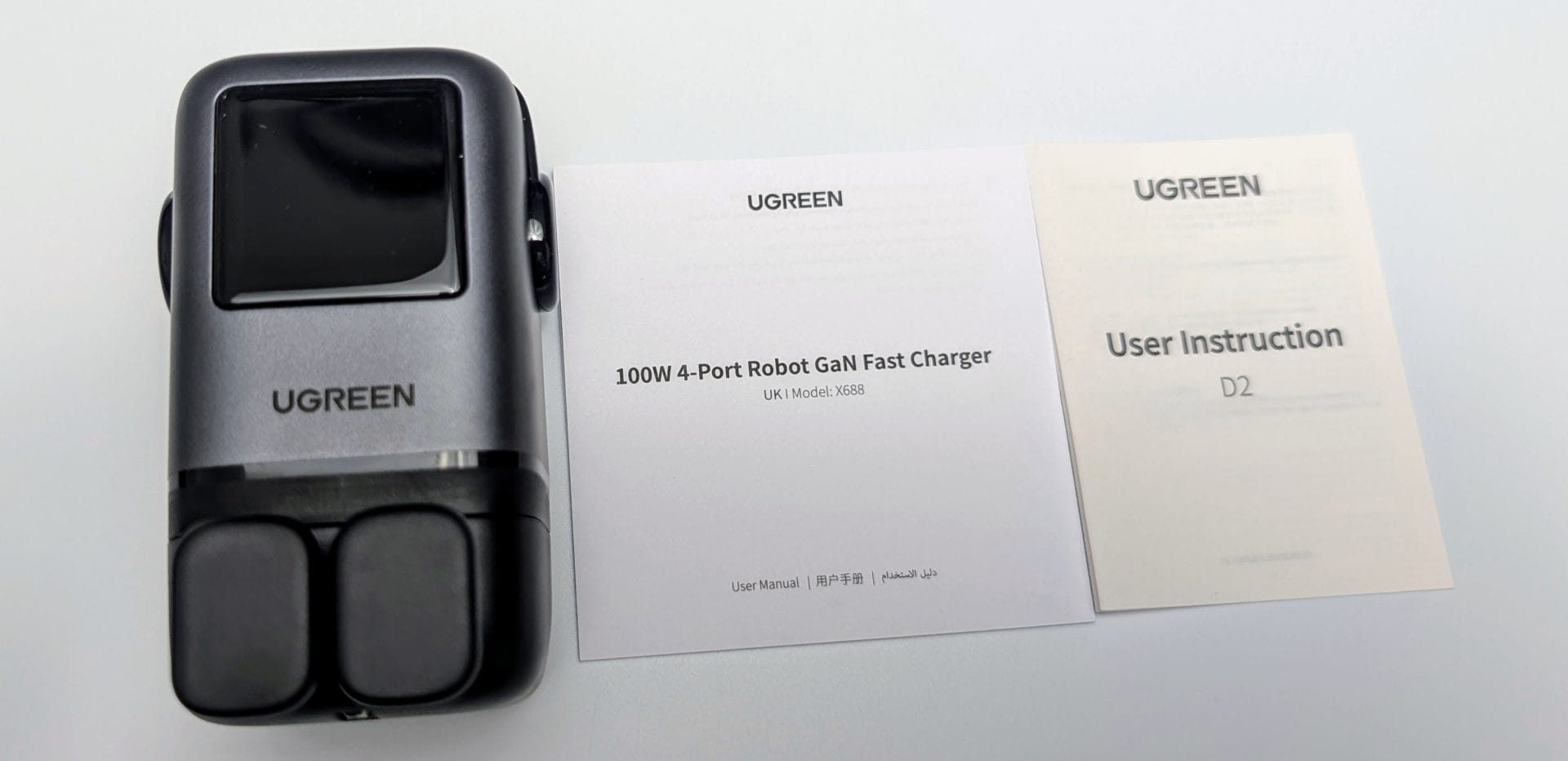 UGREEN Uno Charger 100W A UGREEN 100W 4-Port Robot GaN Fast Charger (UK Model: X688) placed on a white background, featuring a sleek grey and black design with a digital display screen and two charging pads. Next to it are two user manuals, one labeled "100W 4-Port Robot GaN Fast Charger" and the other "User Instruction D2," both with the UGREEN logo.