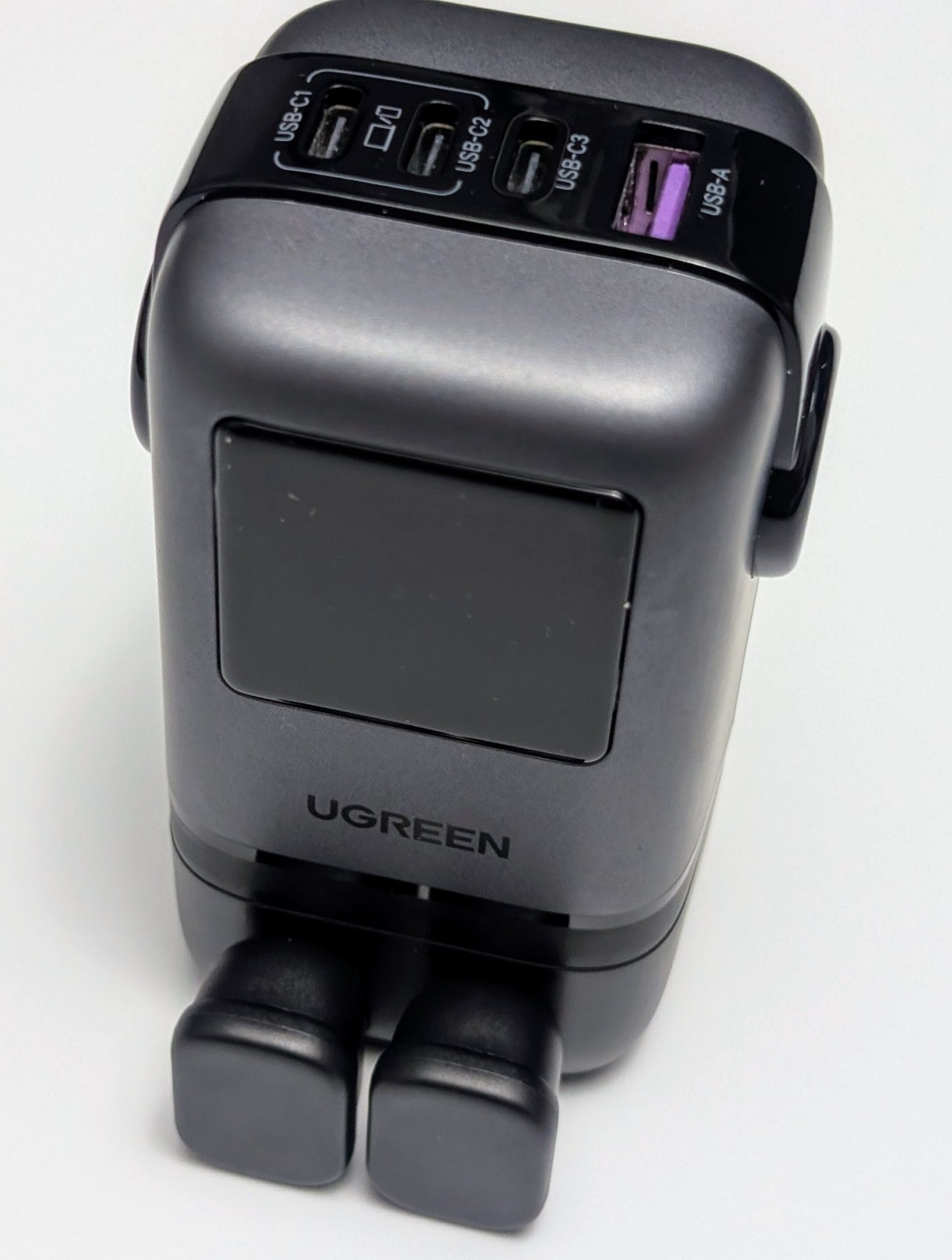UGREEN Uno Charger 100W A UGREEN 100W 4-Port Robot GaN Fast Charger on a white background, featuring a grey and black design with a digital display screen and two black charging pads at the base. The top panel includes four ports: two USB-C ports (labeled USB-C1 and USB-C2/C3) and one USB-A port with a purple insert.