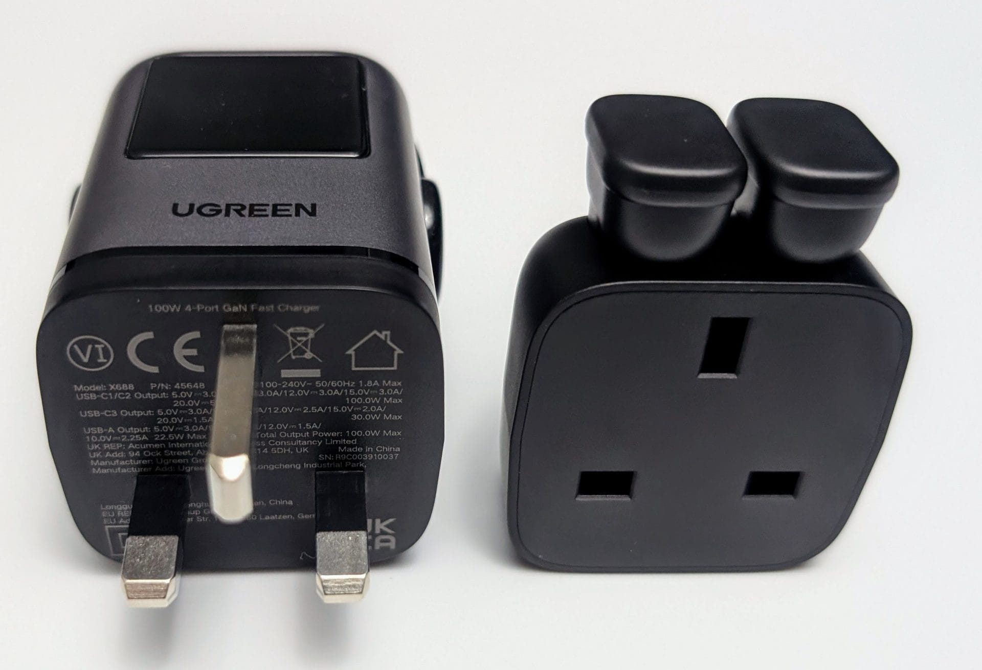 UGREEN Uno Charger 100W A UGREEN 100W 4-Port GaN Fast Charger with a UK plug on a white background, showing the black and grey device with a digital display and two black charging pads. Next to it is a separate black UK plug adapter with three rectangular slots. The charger label includes model details (X688), certifications (CE), and output specifications for USB-C and USB-A ports.