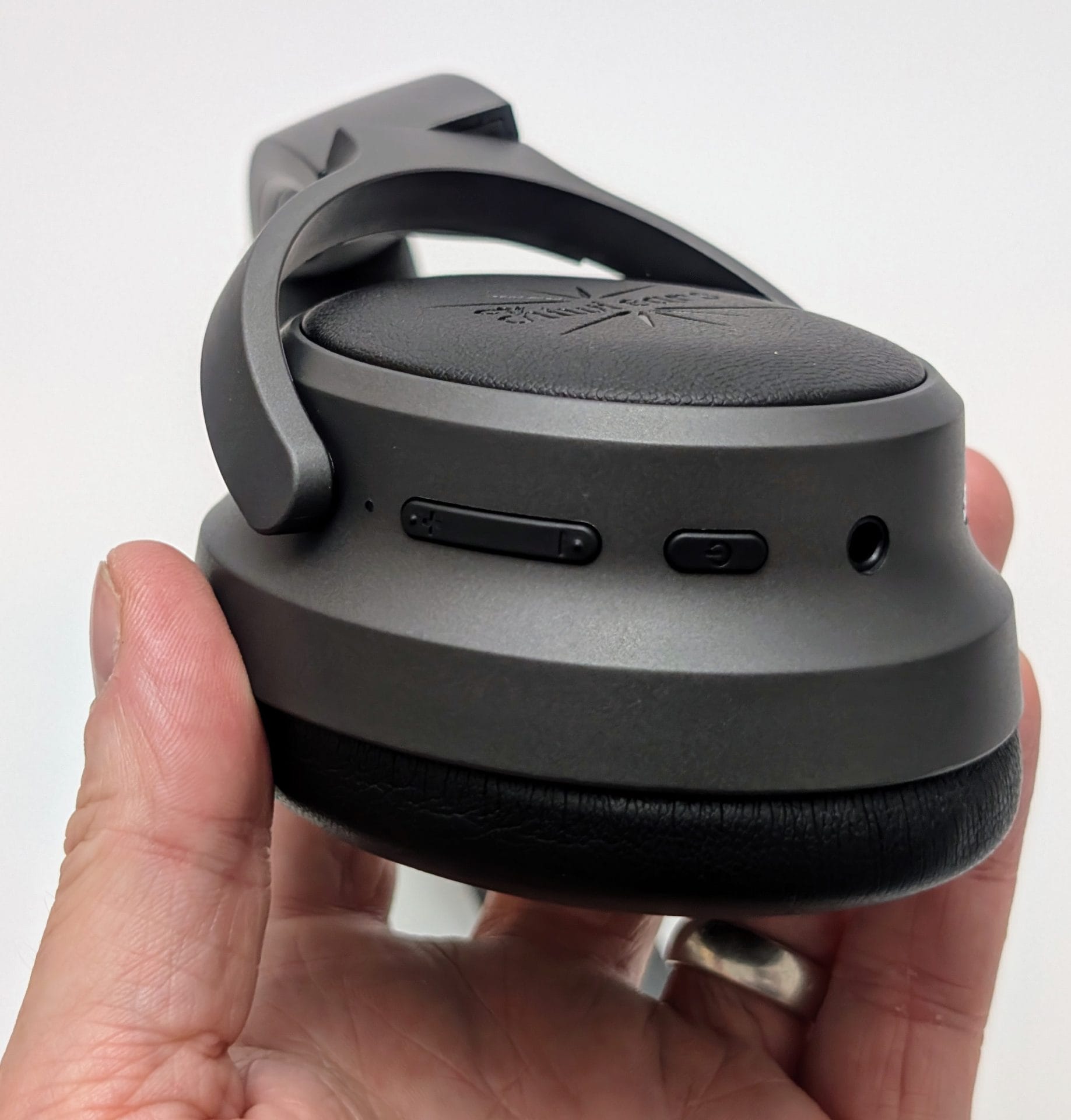 Kiwi Ears Ardor A hand holding a Kiwi Ears Ardor headphone ear cup, showing a black and gray design with the "Kiwi Ears" logo embossed on the leather-like surface. The side features control buttons and a 3.5mm audio jack.