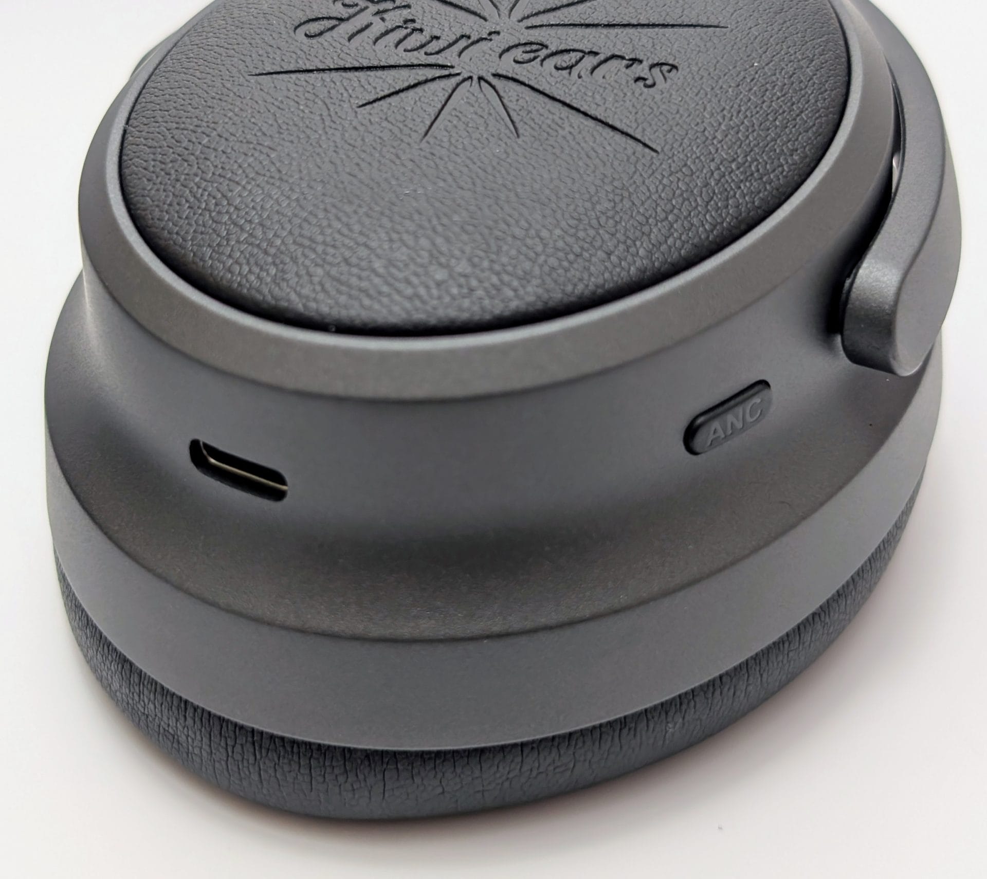 A close-up view of a Kiwi Ears Ardor headphone ear cup, showing a black leather-like surface with the "Kiwi Ears" logo embossed on it. The ear cup features a USB-C port and an "ANC" (Active Noise Cancellation) button on the side.