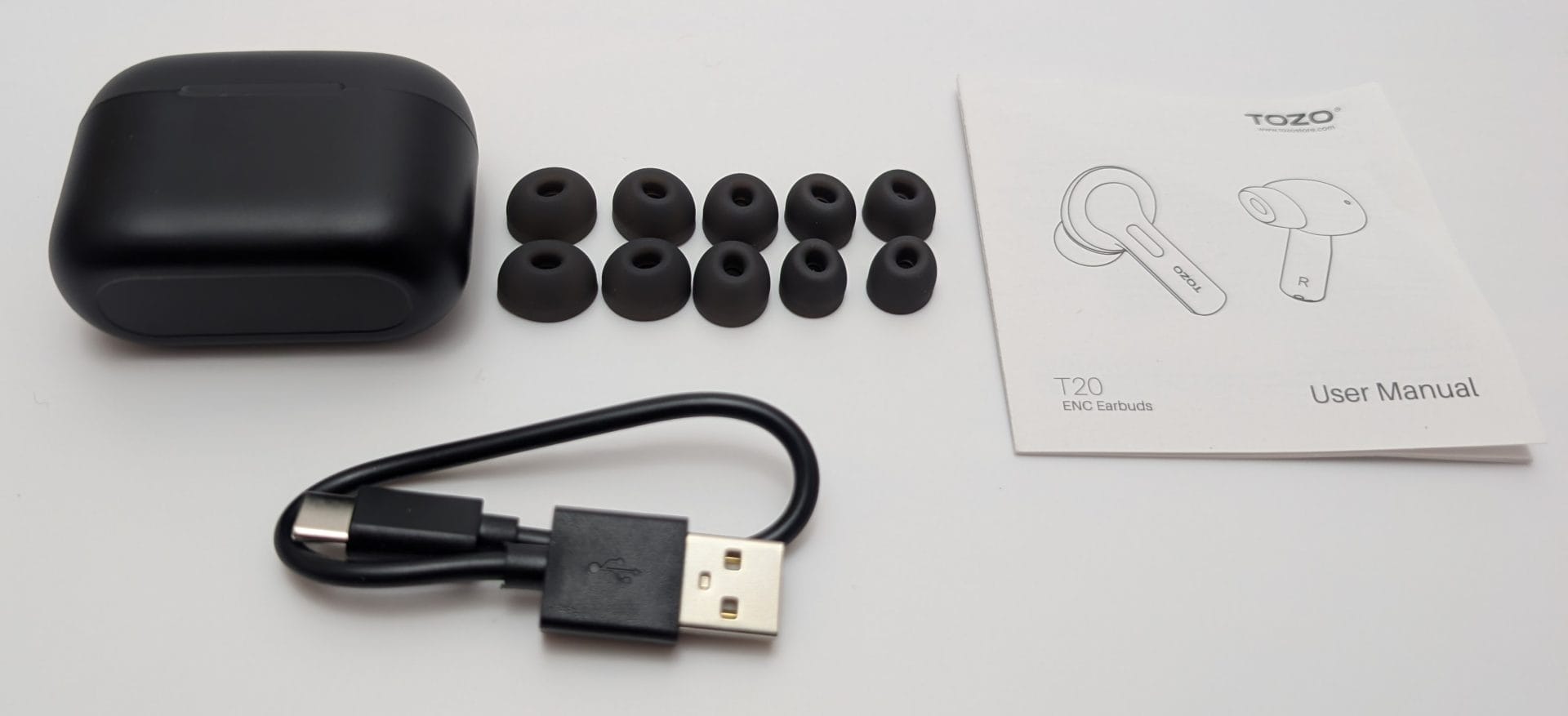 Image of TOZO T20 earbuds accessories, including a black charging case, a USB-A to USB-C charging cable, multiple pairs of black ear tips in various sizes, and a user manual with diagrams of the earbuds labeled "L" and "R".