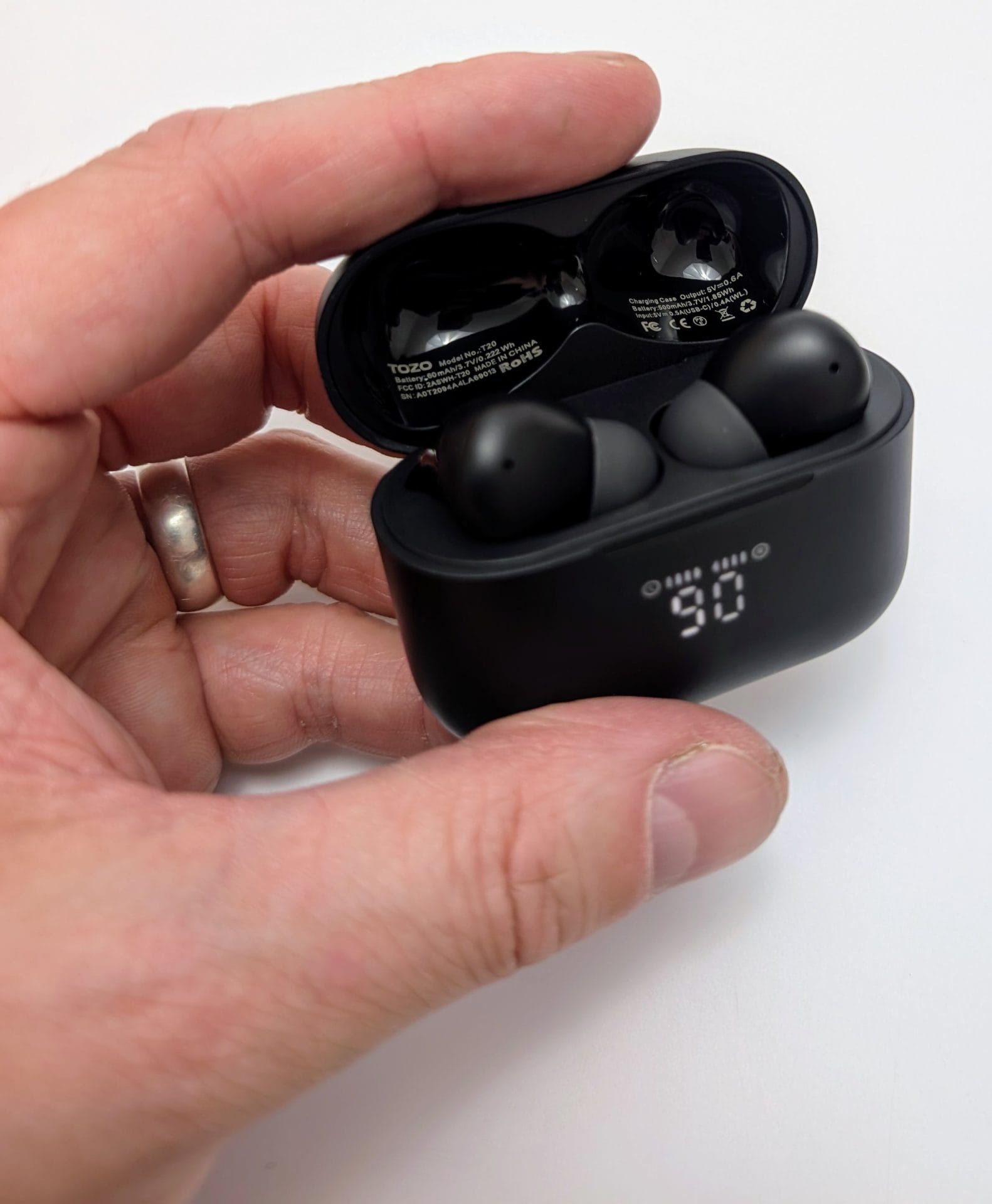 Image of a hand holding an open black TOZO T20 wireless earbuds charging case, displaying a digital battery level of 90%. Two black earbuds are inside the case, with the brand name "TOZO" visible on the interior. The hand is wearing a silver ring.