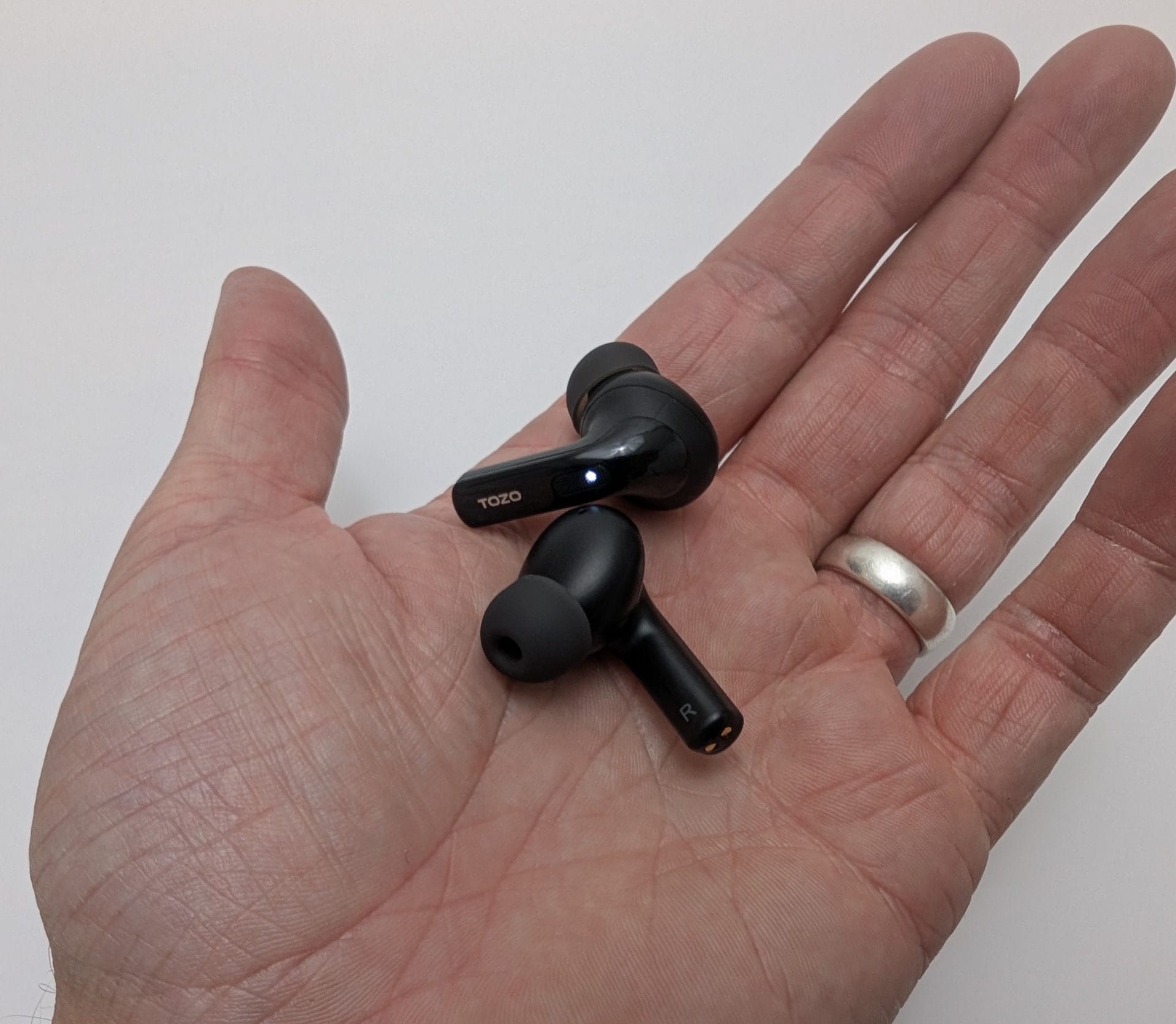Image of a hand holding two black TOZO T20 wireless earbuds, one with a blue light indicator and labeled "L," the other labeled "R," against a plain white background. The hand is wearing a silver ring on the ring finger.