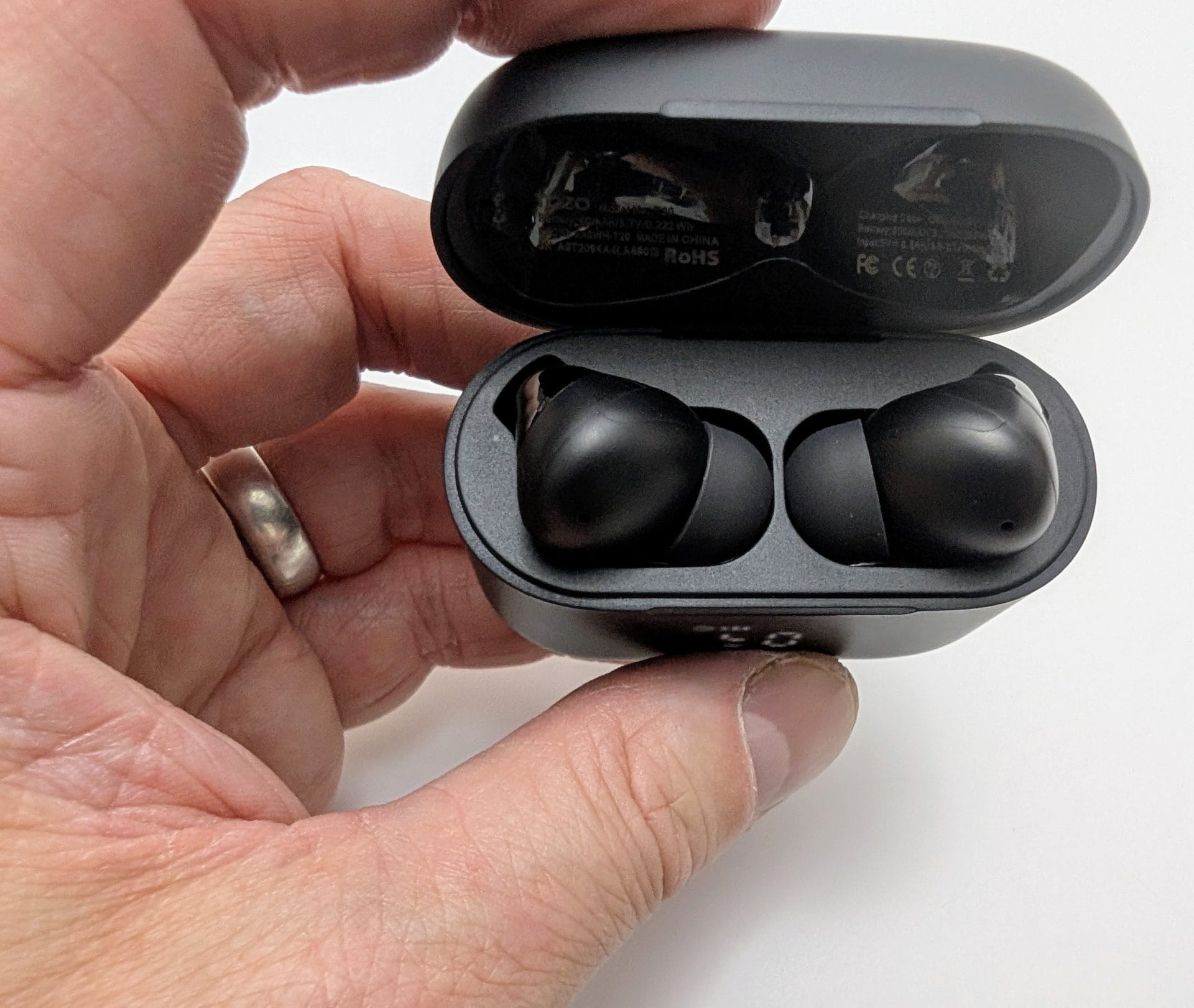 TOZO T20 Wireless Earbuds Image of a hand holding an open black TOZO T20 wireless earbuds charging case, displaying two black earbuds inside. The case shows a digital battery level of 50% on the front, with certification labels visible on the interior lid, against a white background. The hand is wearing a silver ring.