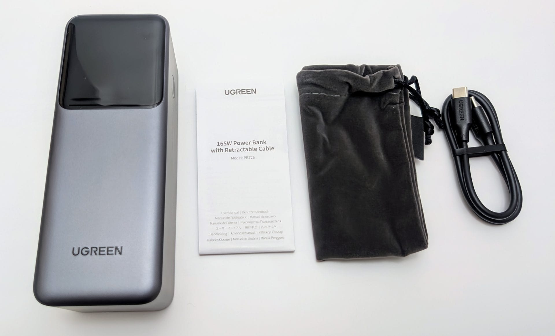 UGREEN Nexode 20000mAh 165W Power Bank with Retractable USB-C Cable unboxed. The image shows the sleek silver and black power bank, a user manual, a gray velvet pouch, and an additional black USB-C cable. The power bank features the UGREEN logo and a glossy top panel.