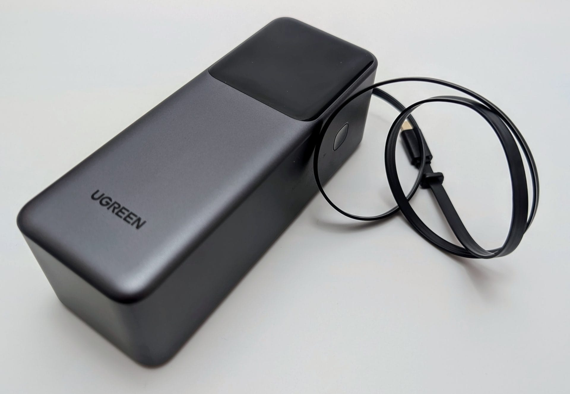 UGREEN Nexode 20000mAh 165W Power Bank with Retractable USB-C Cable. The image shows a sleek, silver and black power bank with a glossy top panel and the UGREEN logo. A retractable black USB-C cable is extended from the device, coiled neatly beside it, on a white background.