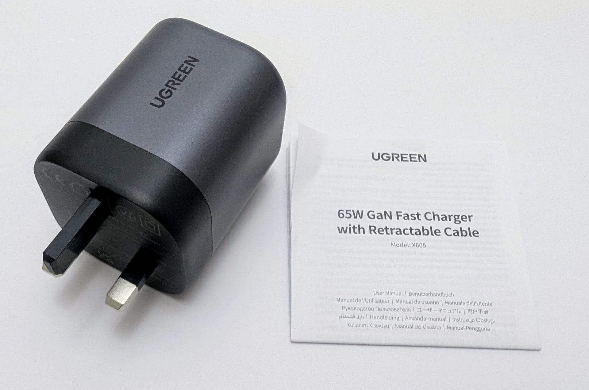 UGREEN Nexode 65W Charger with Retractable USB-C Cable UGREEN 65W GaN Fast Charger with Retractable Cable, shown unpacked on a white surface. The charger is a sleek, black and gray device with a UK plug and the "UGREEN" logo on the side. Next to it lies a user manual titled "65W GaN Fast Charger with Retractable Cable," with model number X605, available in multiple languages.