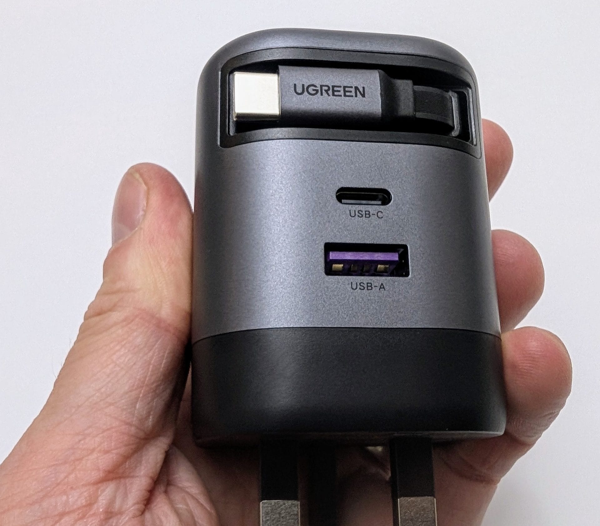 UGREEN Nexode 65W Charger with Retractable USB-C Cable A hand holding a UGREEN charger with a retractable USB-C cable. The charger is gray and black with a UK plug at the bottom. It features two ports: a USB-C port and a USB-A port with a purple interior. The retractable cable, branded with "UGREEN," is partially extended from the top of the charger.