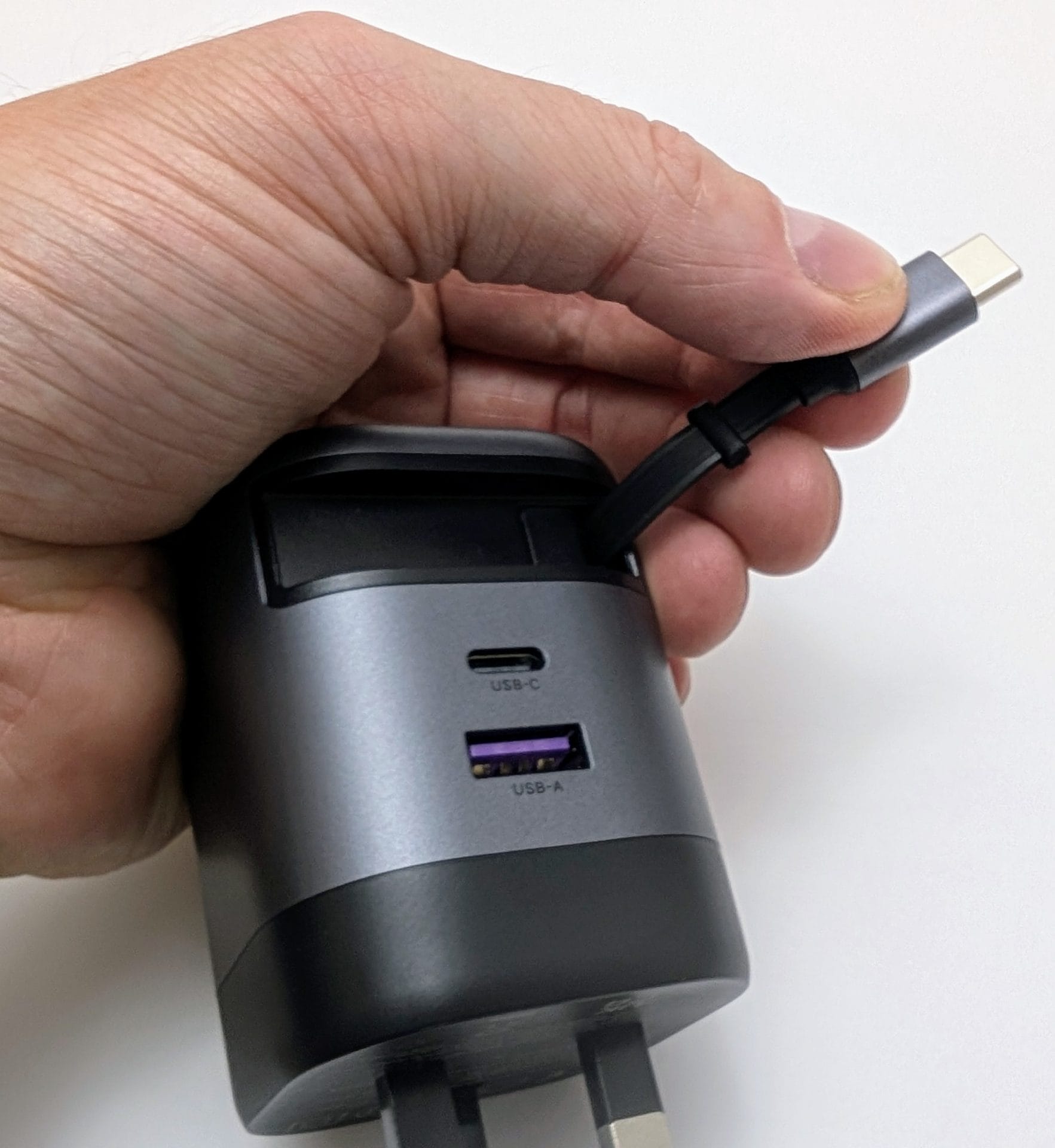 UGREEN Nexode 65W Charger with Retractable USB-C Cable A hand holding a UGREEN charger with a retractable USB-C cable being pulled out. The charger is gray and black with a UK plug at the bottom. It features two ports: a USB-C port and a USB-A port with a purple interior. The cable is partially extended, showing the USB-C connector.