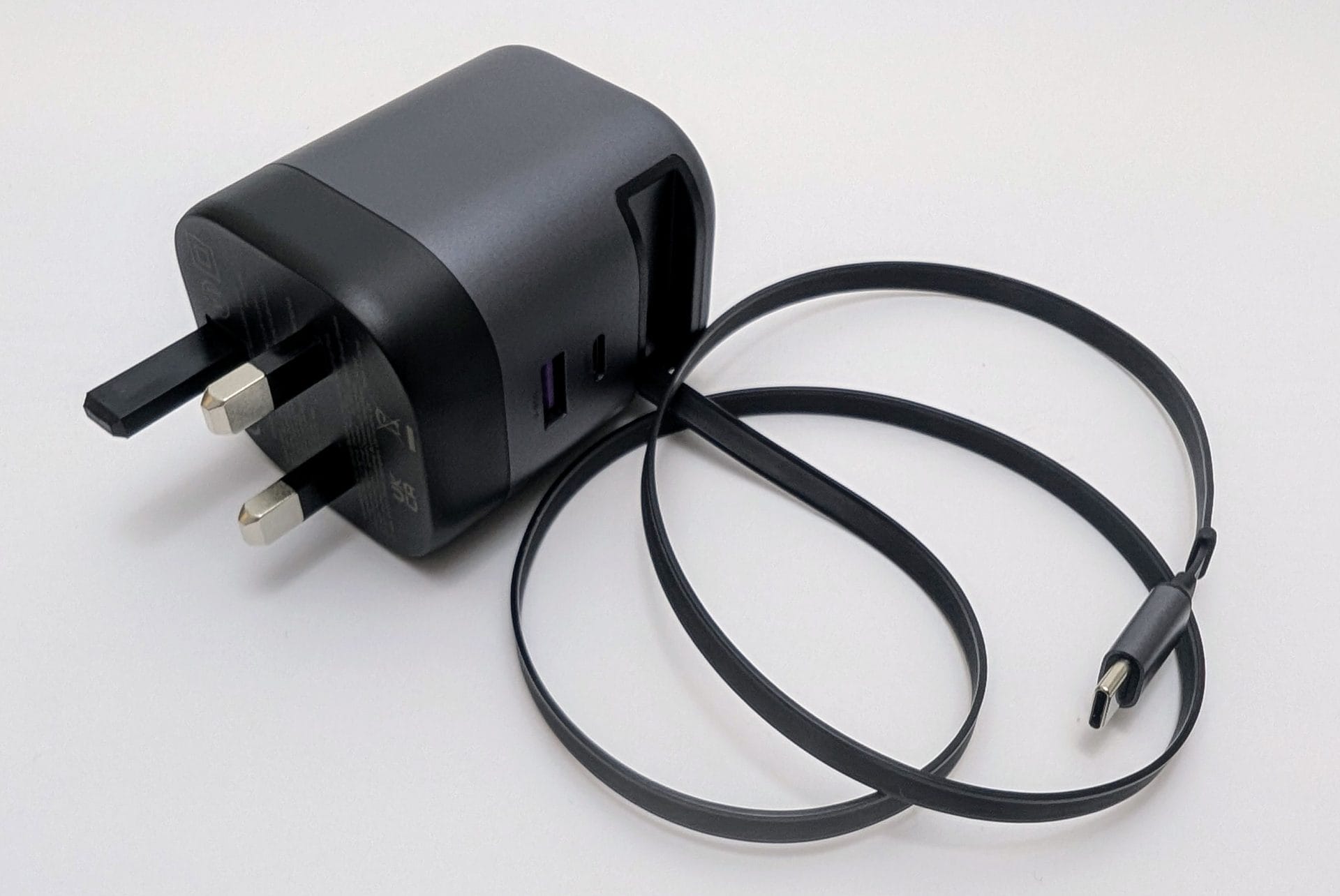 A UGREEN charger with a retractable USB-C cable placed on a white surface. The charger is gray and black with a UK plug at the bottom and two ports (USB-C and USB-A with a purple interior). The black retractable cable is fully extended, coiled loosely, with the USB-C connector visible at the end.