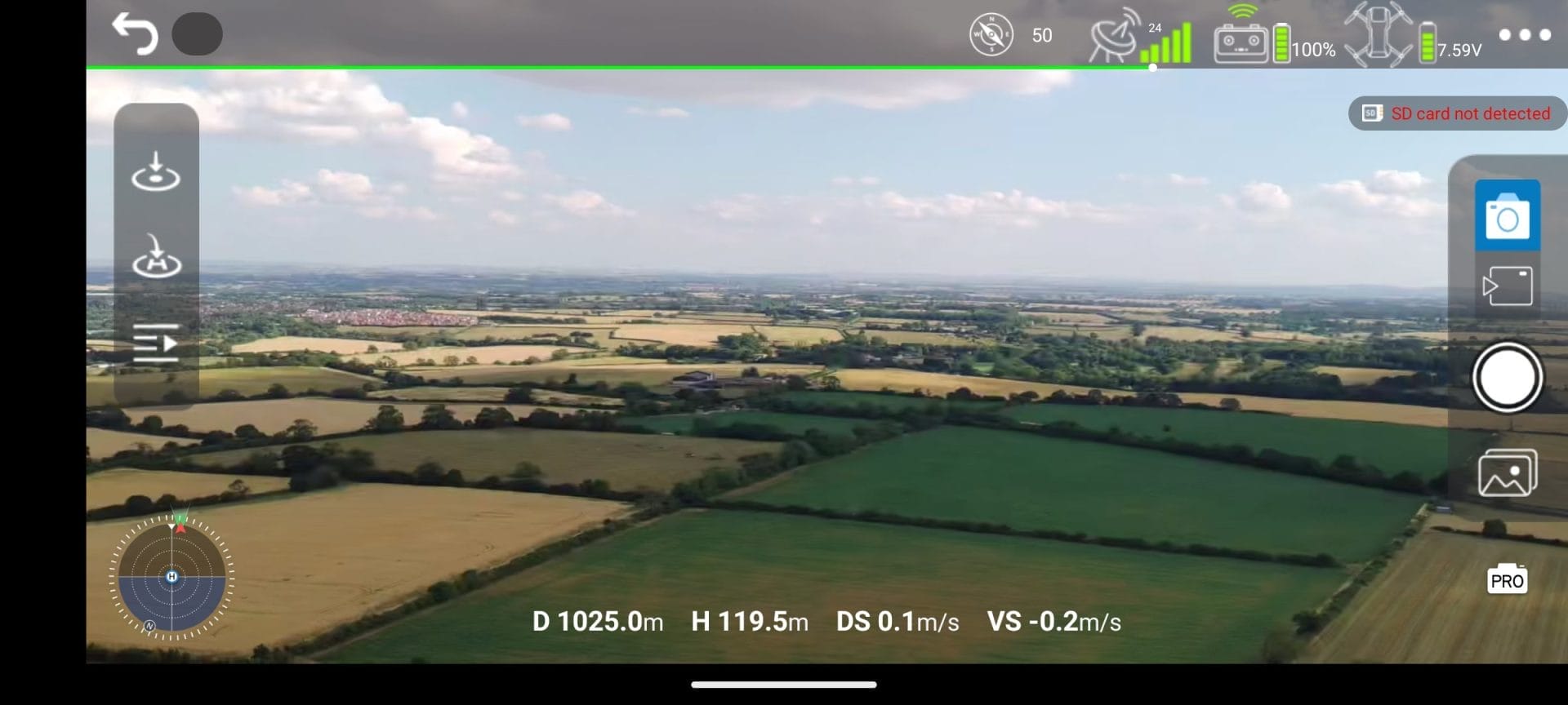 Aerial view from a Ruko U11MINI 4K Drone showing a vast landscape of green and golden fields, scattered trees, and distant villages under a partly cloudy sky. The drone's interface displays a distance of 1025.0m, height of 119.5m, descent speed of 0.1m/s, and vertical speed of -0.2m/s. The battery is at 100% with 7.59V, and an SD card is not detected. On-screen controls include navigation, camera, and photo options.