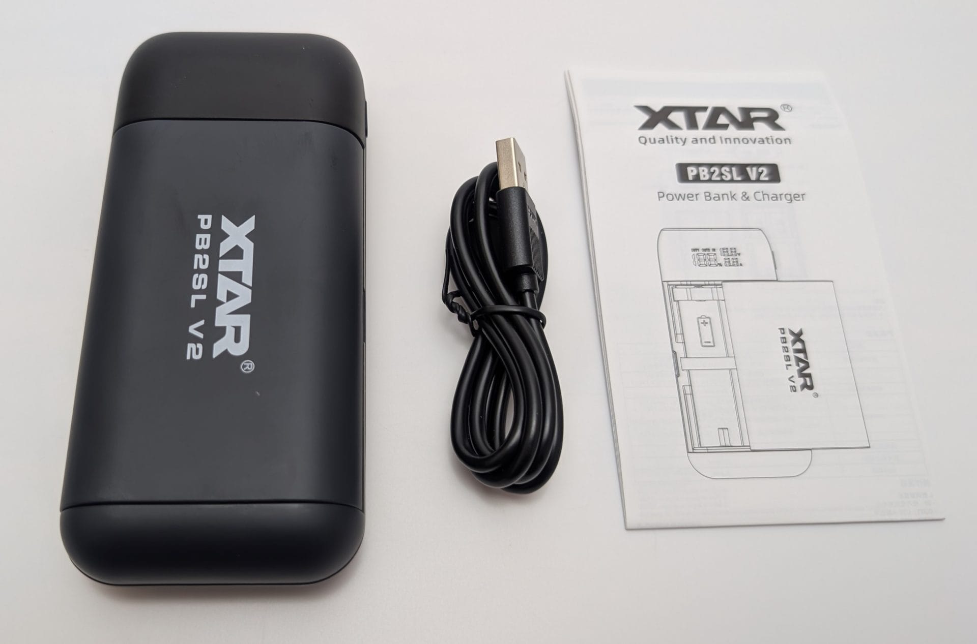 XTAR PB2SL V2 Alt text: Unboxed XTAR PB2SL V2 power bank and charger in black, displayed with a coiled USB cable and a user manual. The manual, branded with XTAR's "Quality and Innovation" logo, includes diagrams and details about the PB2SL V2 power bank and charger.