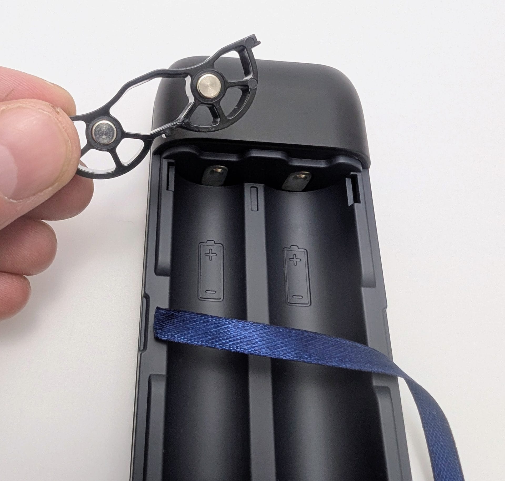 XTAR PB2SL V2 Alt text: Close-up of an XTAR PB2SL V2 power bank with its top cover removed, revealing two battery slots with positive and negative indicators. A hand holds a black plastic component with metal contacts, likely part of the battery holder. A blue ribbon is visible inside the device.