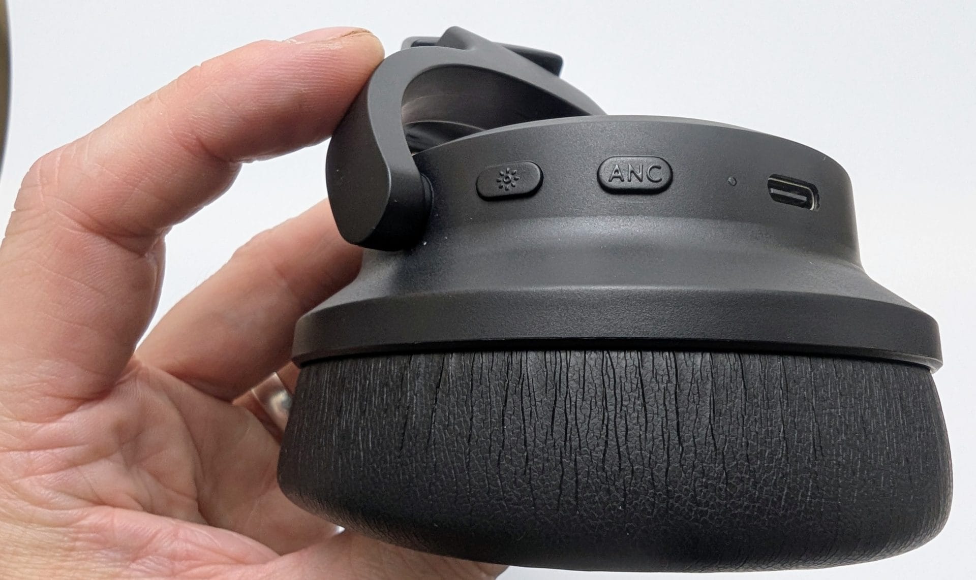 A close-up of a hand holding a black Kiwi Ears Aventus headphone ear cup, displaying the cushioned exterior, a power button with a light icon, an ANC (Active Noise Cancellation) button, and a USB-C port. The background is a plain white surface.