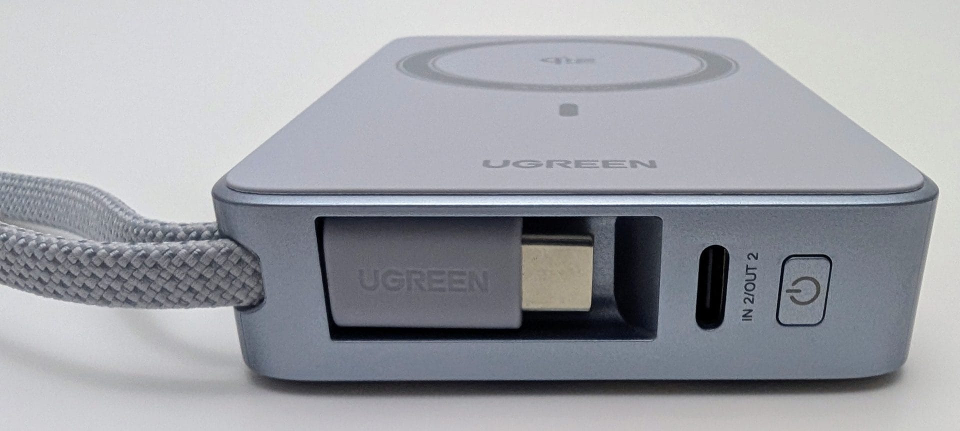 A close-up view of the side of a UGREEN MagFlow 10000mAh Magnetic Wireless Power Bank, showcasing a built-in braided strap, a retractable UGREEN-branded USB-C cable, a USB-C input/output port labeled "IN 2I OUT," and a power button. The device features a circular Qi2 wireless charging pad on top, set against a plain white background.