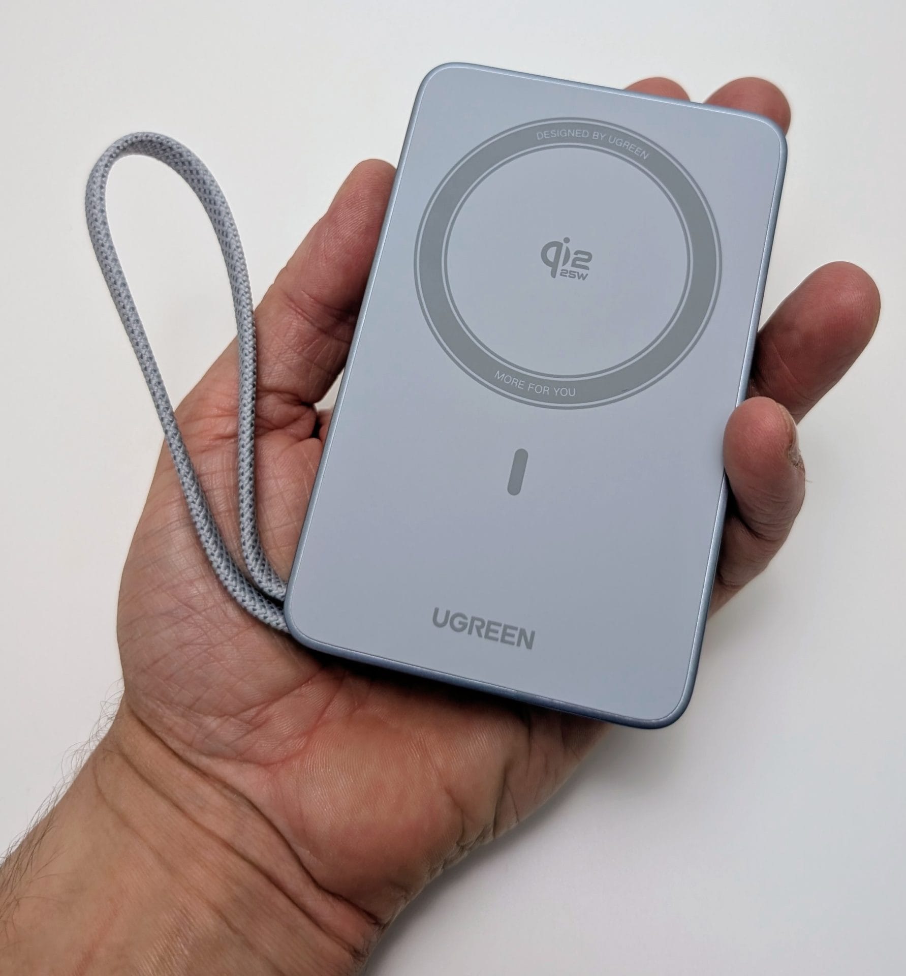 A hand holding a UGREEN MagFlow 10000mAh Magnetic Wireless Power Bank with a 25W Qi2-certified wireless charging pad. The sleek gray device features a circular charging area with the "qi2 25W" logo, a power button, and the "UGREEN" branding. It includes a braided wrist strap for easy carrying, displayed against a plain white background.