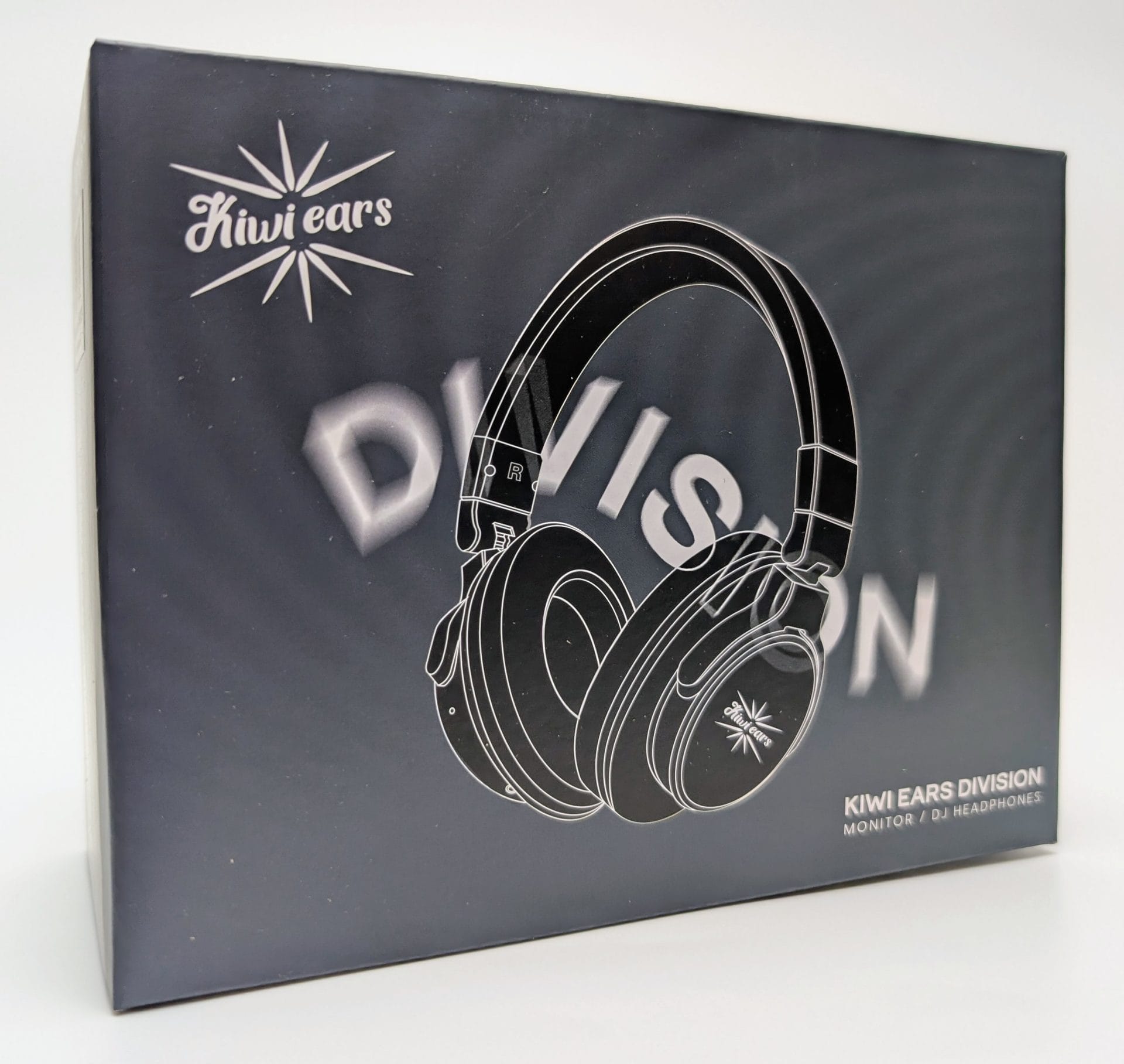 A close-up photograph of the Kiwi Ears Division headphone packaging box. The box has a matte black finish with a stylized white outline illustration of over-ear headphones centered on the front. The Kiwi Ears logo (a stylized starburst) appears in the top left and on the right earcup of the illustrated headphones. Large blurred text “DIVISION” is visible behind the headphones, while smaller clear text in the lower right reads “KIWI EARS DIVISION MONITOR / DJ HEADPHONES” in white. The overall design is minimalist and premium-looking.