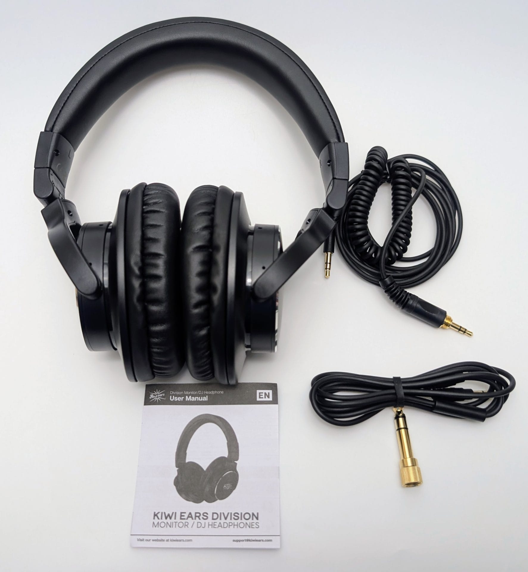 A clean top-down unboxing photo of the Kiwi Ears Division monitor/DJ headphones and included accessories on a white background. The matte black over-ear headphones with soft leather earpads and headband are folded compactly in the center. To the right are two detachable cables: a short coiled cable with a gold-plated 3.5 mm plug and a longer straight cable with a gold-plated 6.35 mm (1/4") adapter attached. Below is the folded user manual featuring the Kiwi Ears logo and text “KIWI EARS DIVISION MONITOR / DJ HEADPHONES” along with a silhouette of the headphones. The overall presentation is minimal and professional.