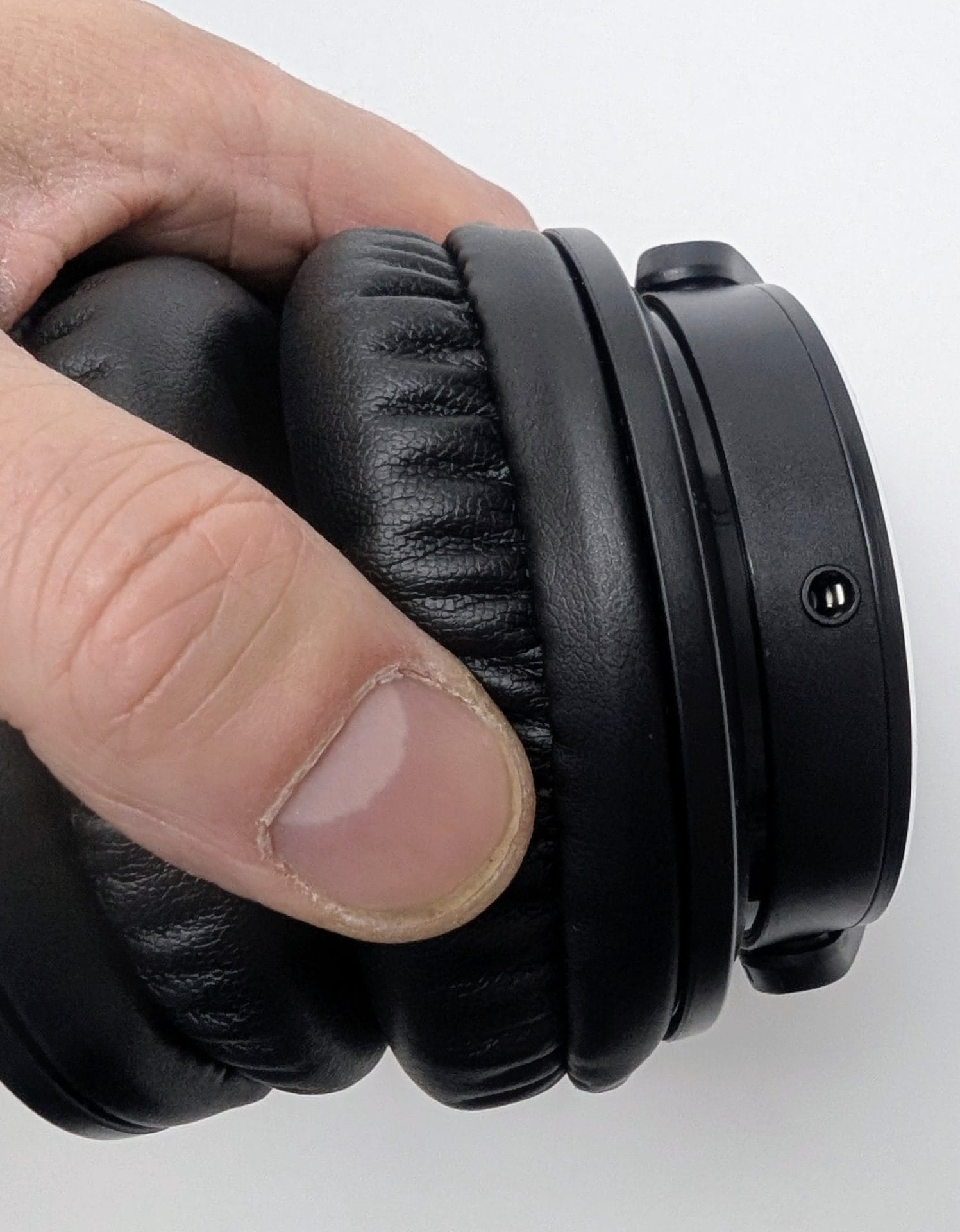 Close-up macro shot of a finger pressing into the thick, plush protein-leather earpad of the Kiwi Ears Division headphones. The pad compresses deeply under light pressure, revealing excellent cushioning and softness, with visible stitched detailing and a small vent port on the matte black plastic housing nearby. Demonstrates the high level of comfort designed for extended listening sessions.