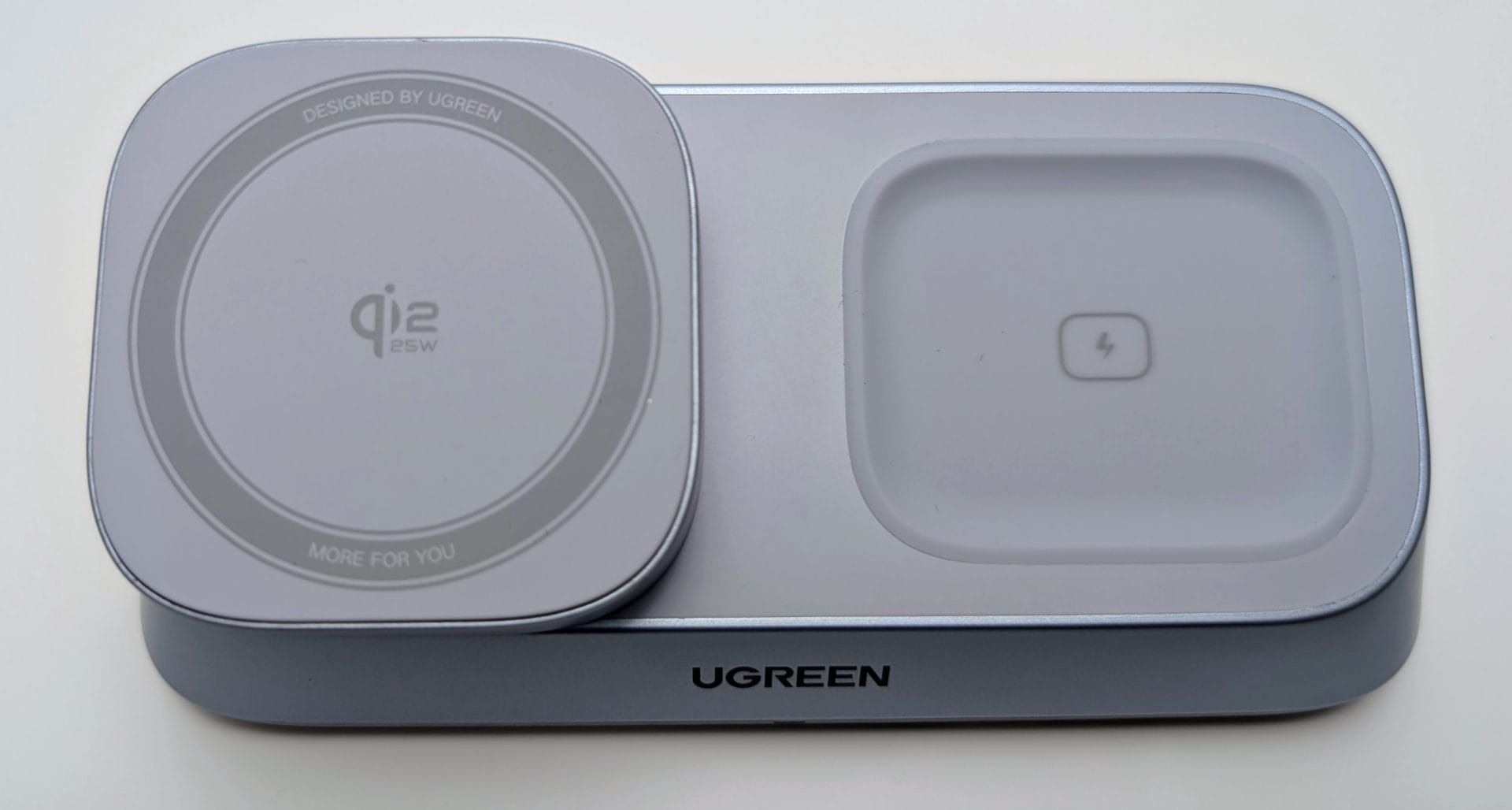 UGREEN MagFlow 3-in-1 Magnetic Wireless Desktop Charger A close-up view of the UGREEN MagFlow 3-in-1 Magnetic Wireless Charger base unit. The grey/light blue charger is shown in its folded-down configuration. The left side features a round, slightly raised MagSafe-compatible charging pad labeled "Qi2 25W" with a concentric ring design. The right side is a recessed, rectangular charging area for AirPods, marked with a small lightning bolt icon. The UGREEN logo is centered on the front edge of the base.