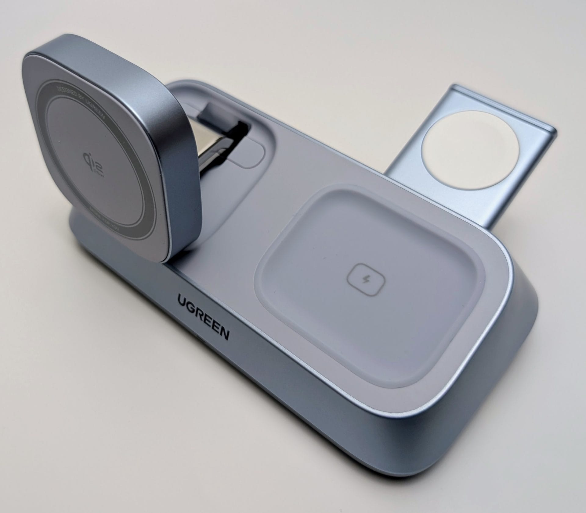 The UGREEN MagFlow 3-in-1 Magnetic Wireless Charger shown in its partially set up state. The main unit is a light blue-grey color. The round, MagSafe-compatible charging pad on the left is angled up and features the Qi2 logo. The separate Apple Watch charging arm is deployed and angled up from the right side, showing its small white magnetic charging surface. The center AirPods charging pad is flat, featuring a lightning bolt icon. The UGREEN logo is visible on the front base.